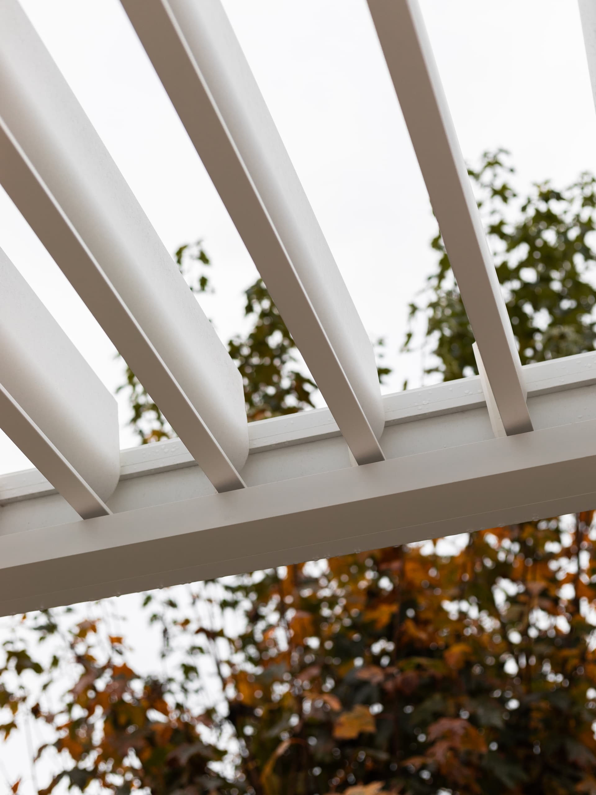 Close‑up van aluminium lamellen van een pergola in gedeeltelijk open stand, met bomen op de achtergrond.