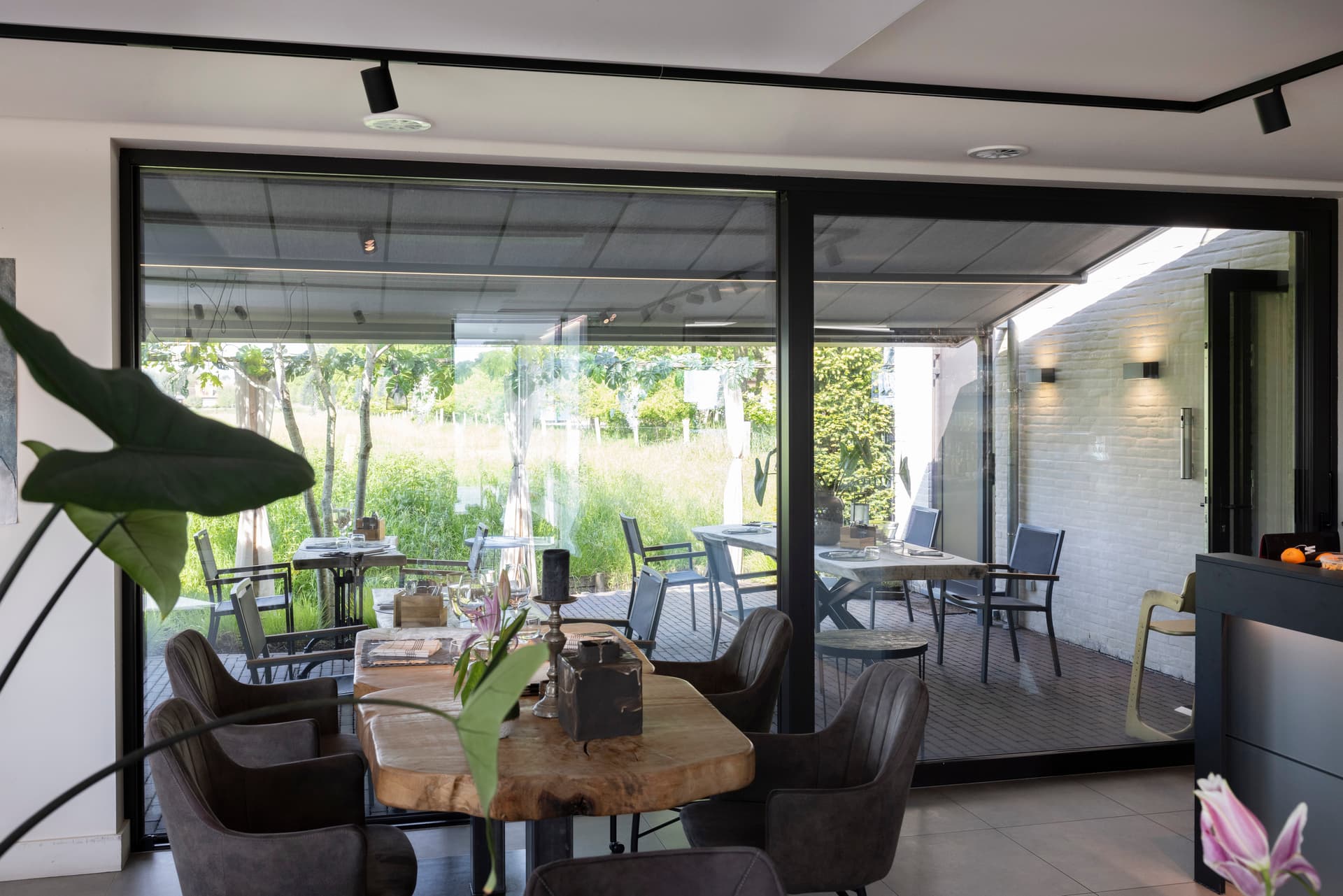 Binnenzicht door grote schuiframen op een aluminium pergola met oprolbaar doekdak boven een moderne buiteneethoek
