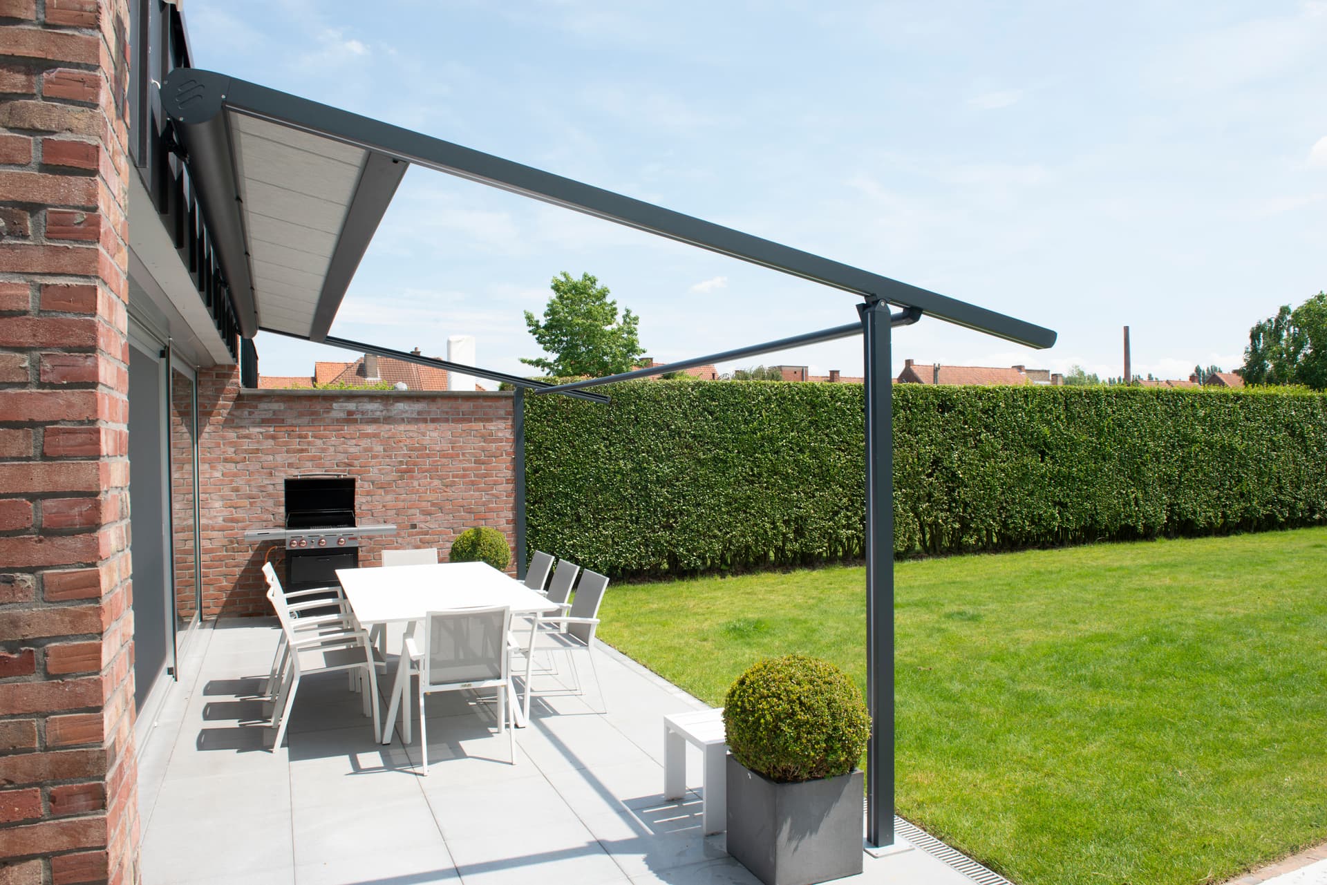 Aluminium pergola met oprolbaar doekdak dat schaduw biedt boven een moderne buiteneettafel naast een bakstenen woning en strakke tuin