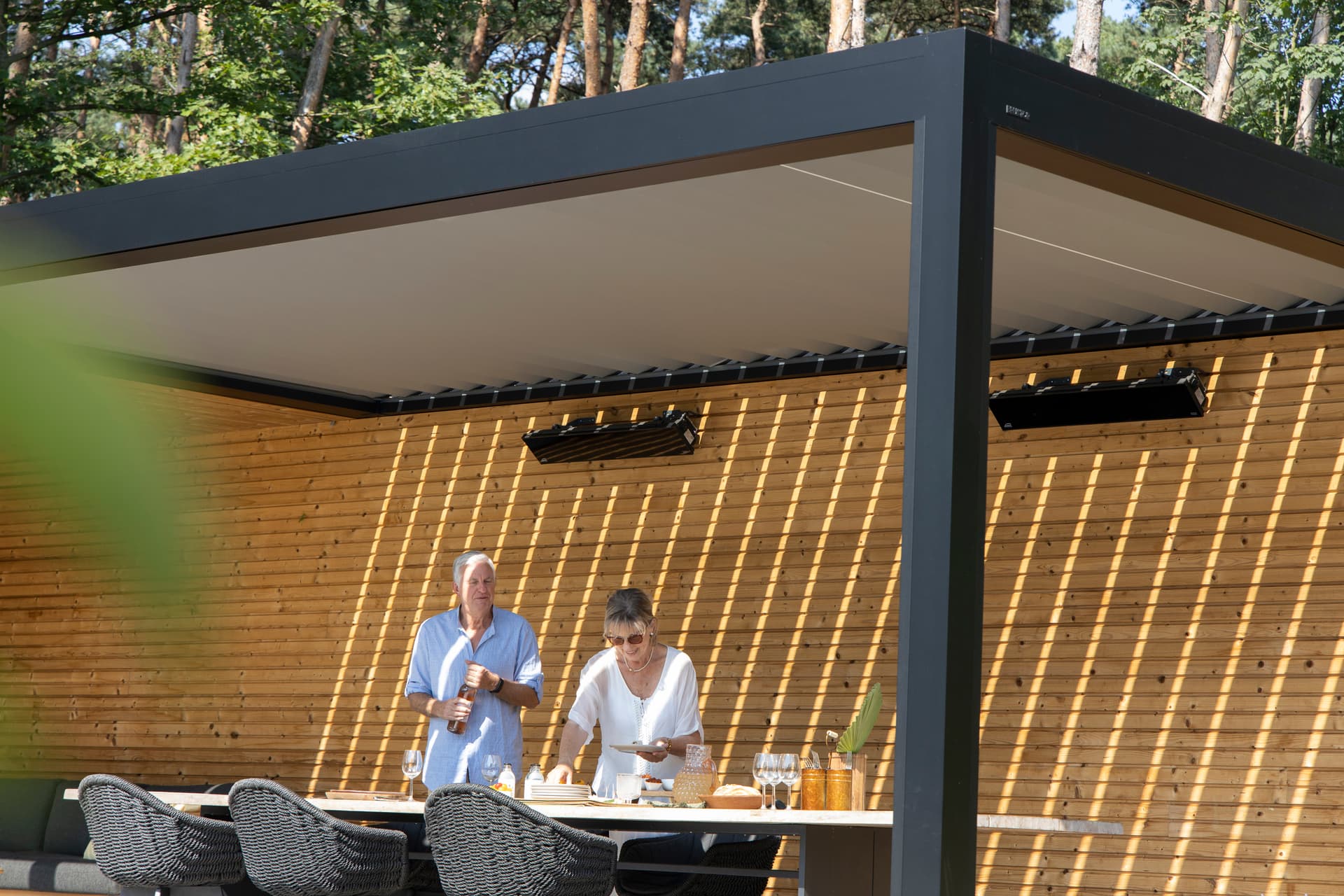 Twee personen die eten voorbereiden aan een buitentafel onder een aluminium pergola met lamellendak en houten lattenwand.