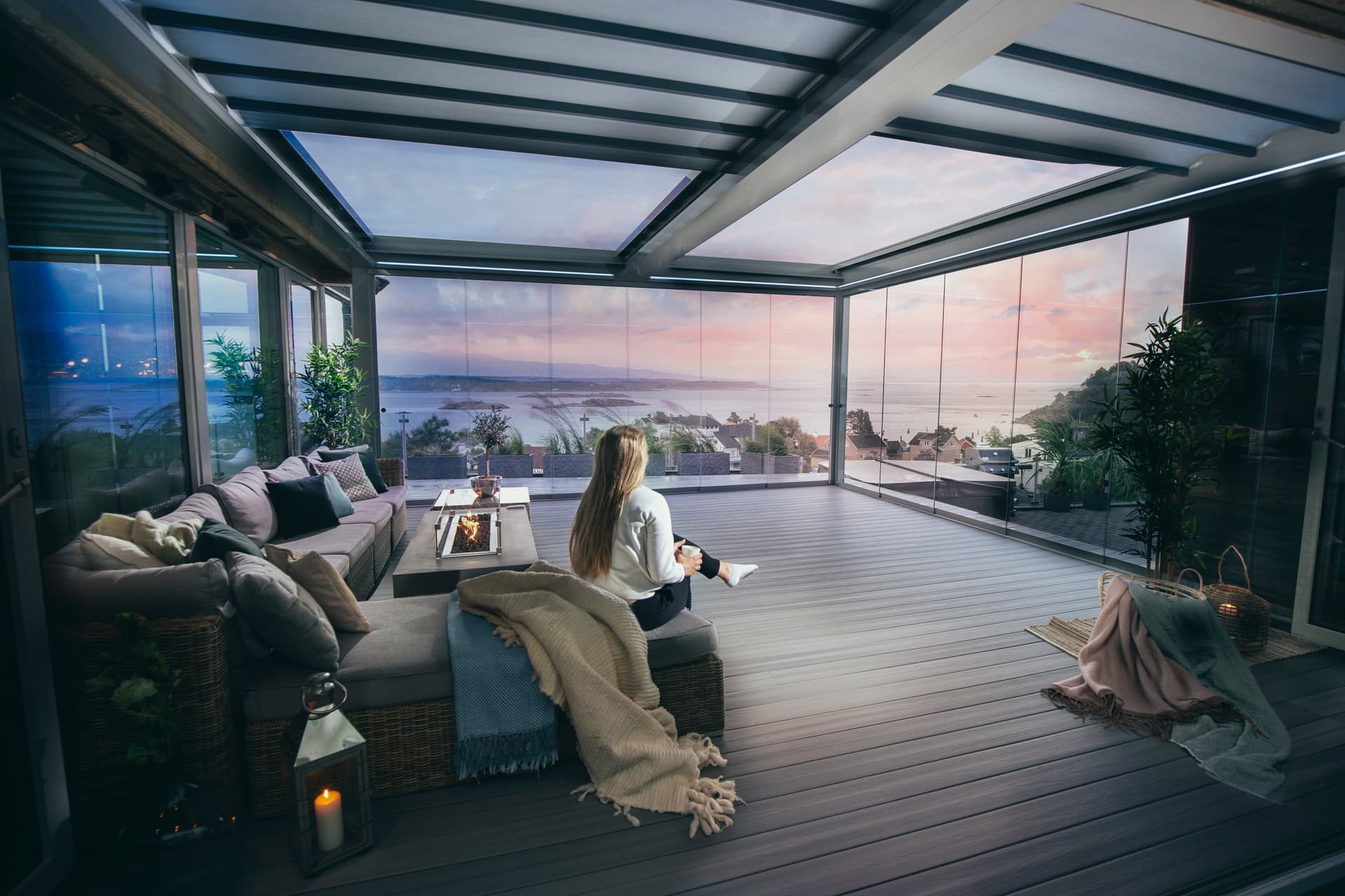 Aluminium pergola met glazen dak en schuifbare glaswanden die een gezellige loungehoek met zeezicht bij zonsondergang vormen