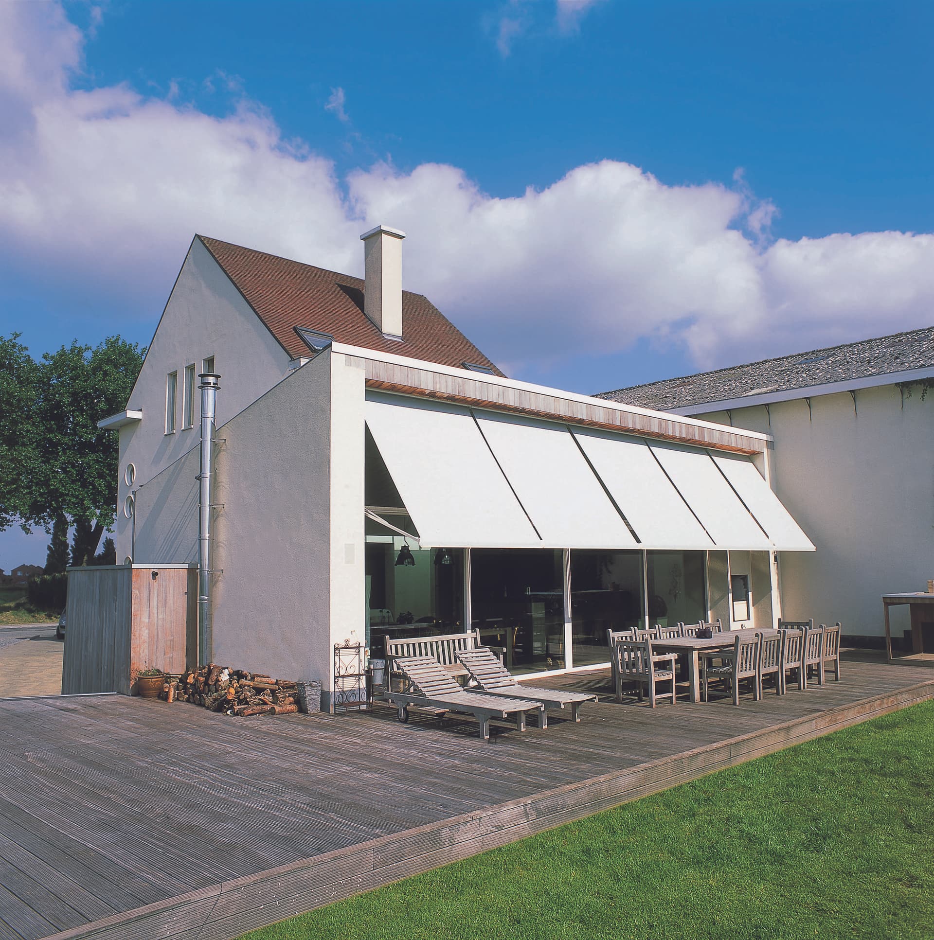 Hellende buitenzonwering die een grote glasgevel en houten terras met lounge‑ en eetmeubilair aan een moderne woning beschaduwt