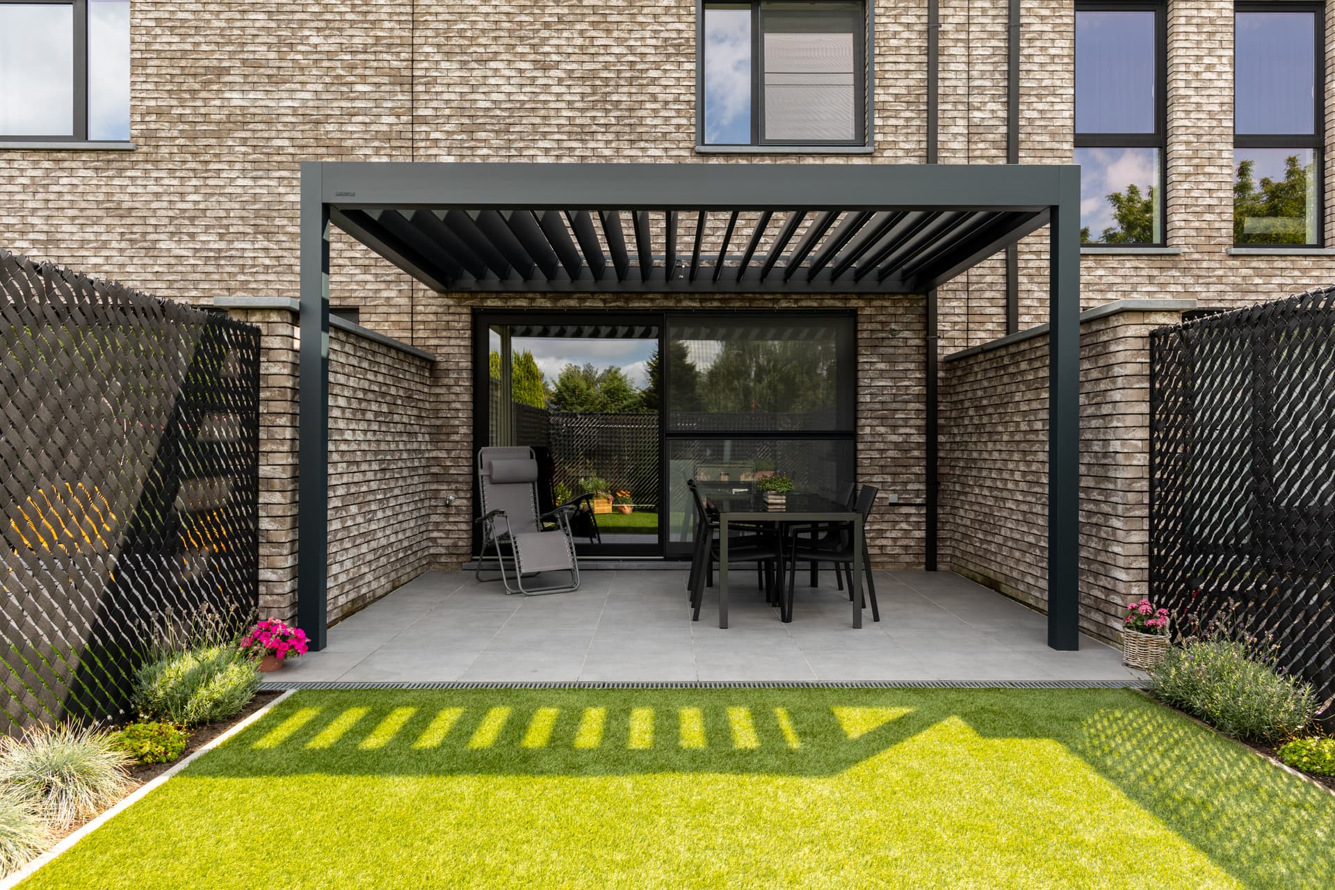 Aluminium pergola met geopend lamellendak dat schuine schaduwpatronen werpt over een terras met buitenmeubilair naast een moderne bakstenen woning.