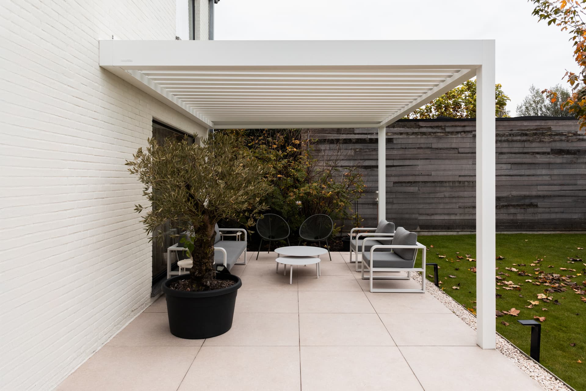 Aluminium pergola met lamellendak op een terras naast een witte bakstenen woning, die een moderne buitenlounge overschaduwt.