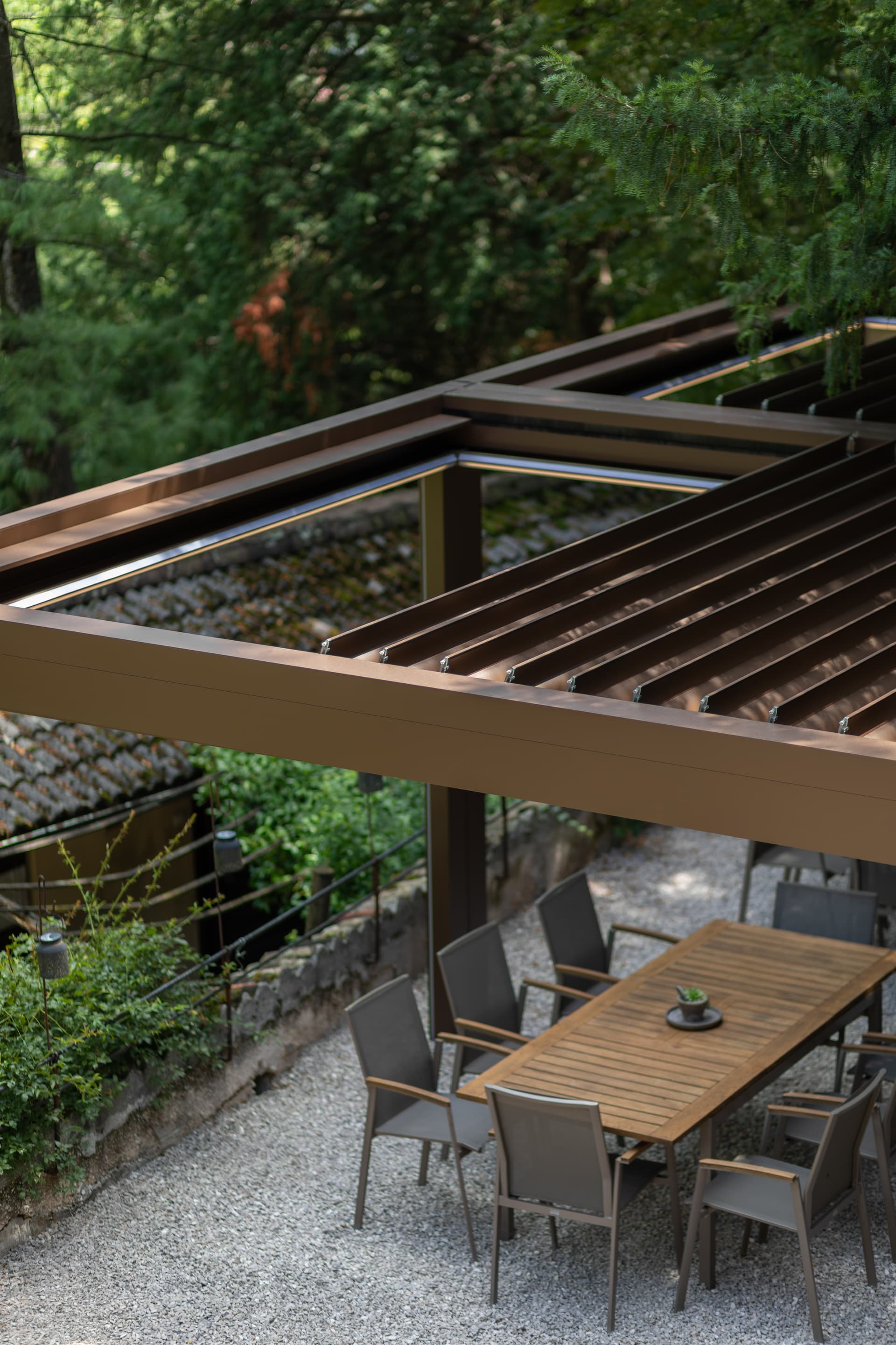 Aluminium terrasoverkapping met openschuifbaar lamellendak in bronzen uitvoering boven een moderne buiteneethoek in groene tuin