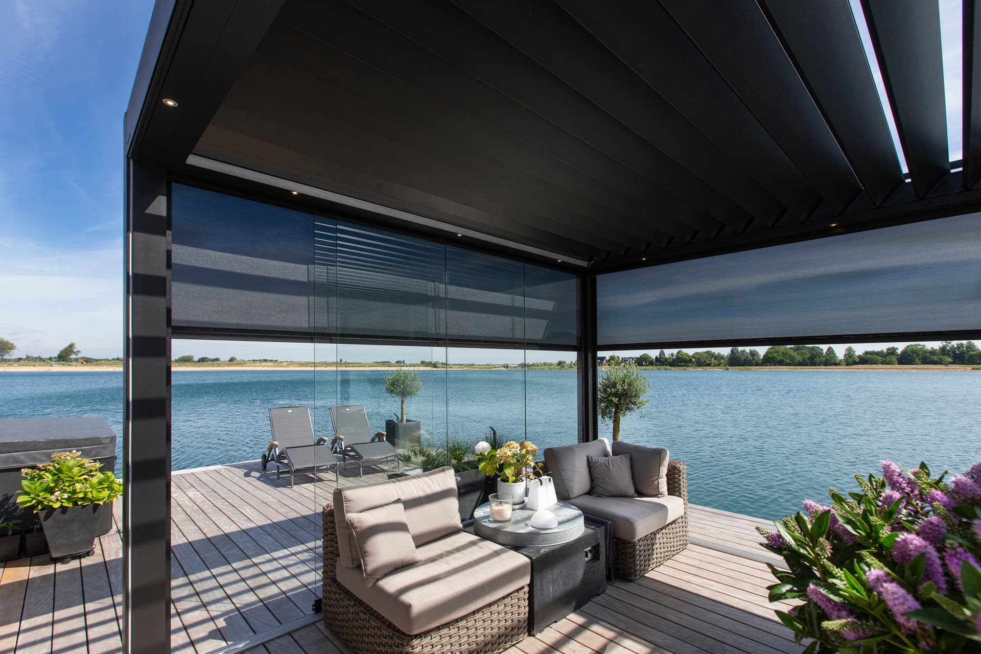 Buitenlounge onder een aluminium pergola met ZIP‑screens, met uitzicht op een waterrijk terras met ligstoelen.