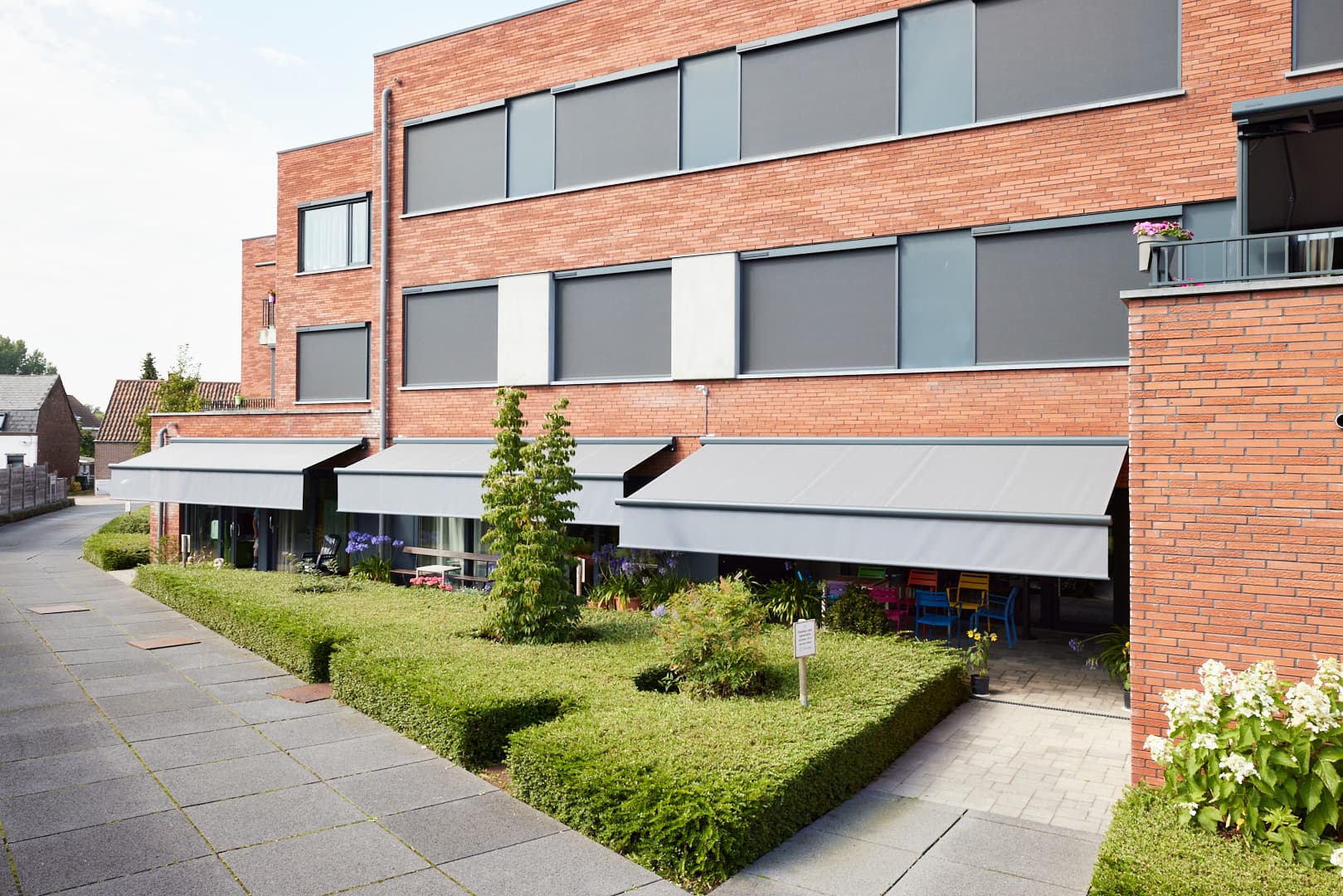 Combinatie van Brustor ZIP‑screens op de bovenverdieping en knikarmschermen op het gelijkvloers die een modern bakstenen gebouw met binnentuin beschaduwen