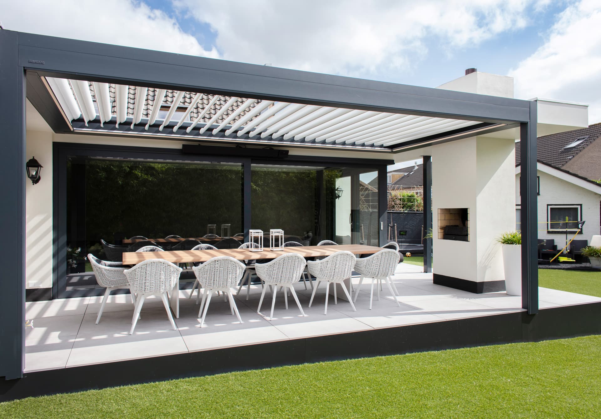 Aluminium terrasoverkapping met openschuifbaar lamellendak boven een ruime buiteneettafel met ingebouwde haard in een moderne tuin