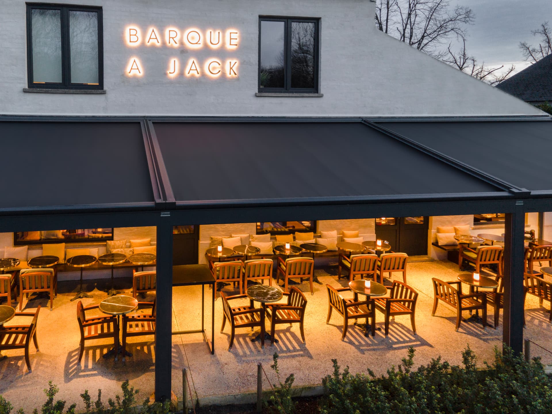 Aluminium B128 XL‑pergola met volledig uitgerold doekdak boven een sfeervol restaurantterras met warme verlichting en houten buitenmeubilair