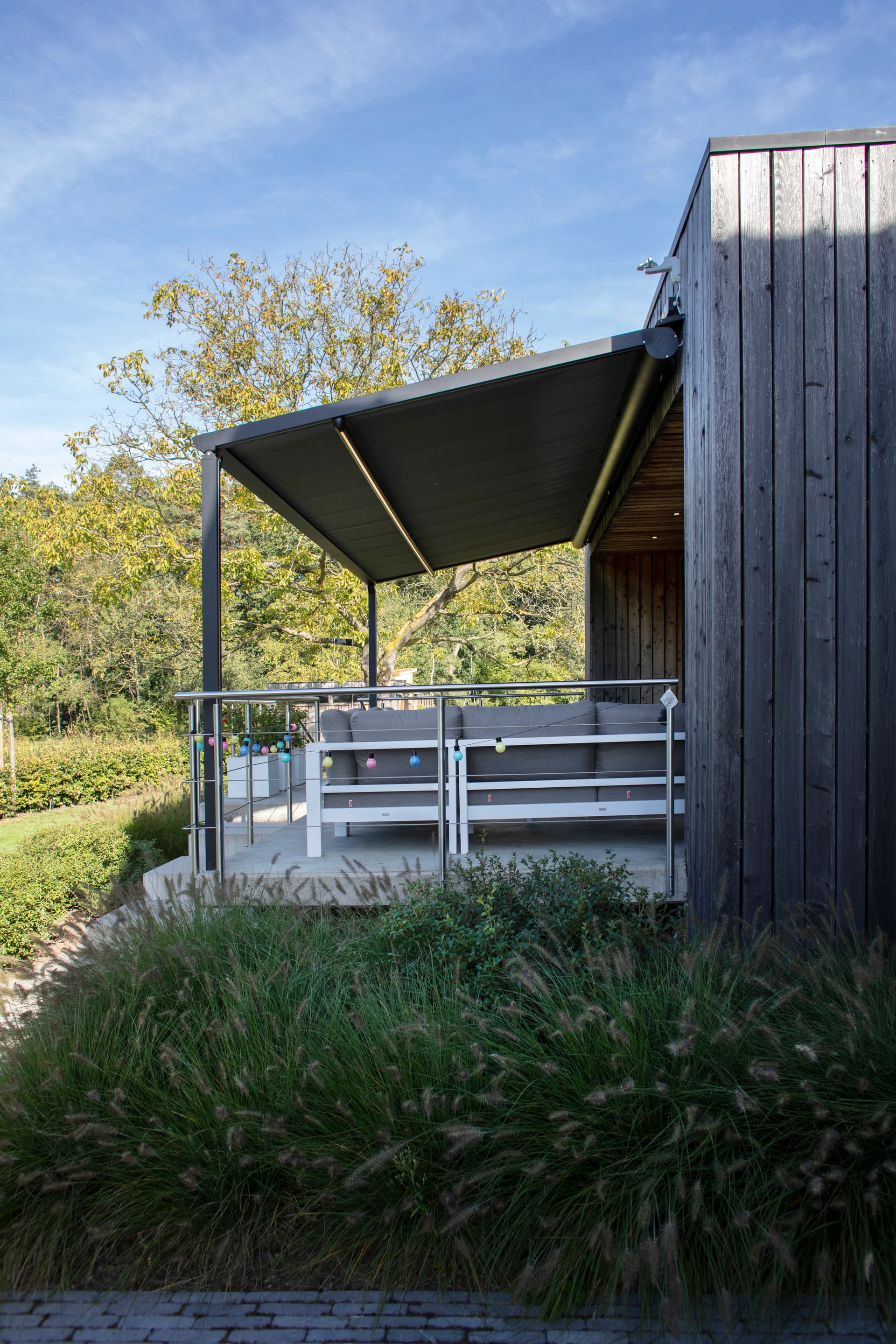 Aluminium pergola met oprolbaar doekdak boven een verhoogde loungehoek aan een moderne houten woning