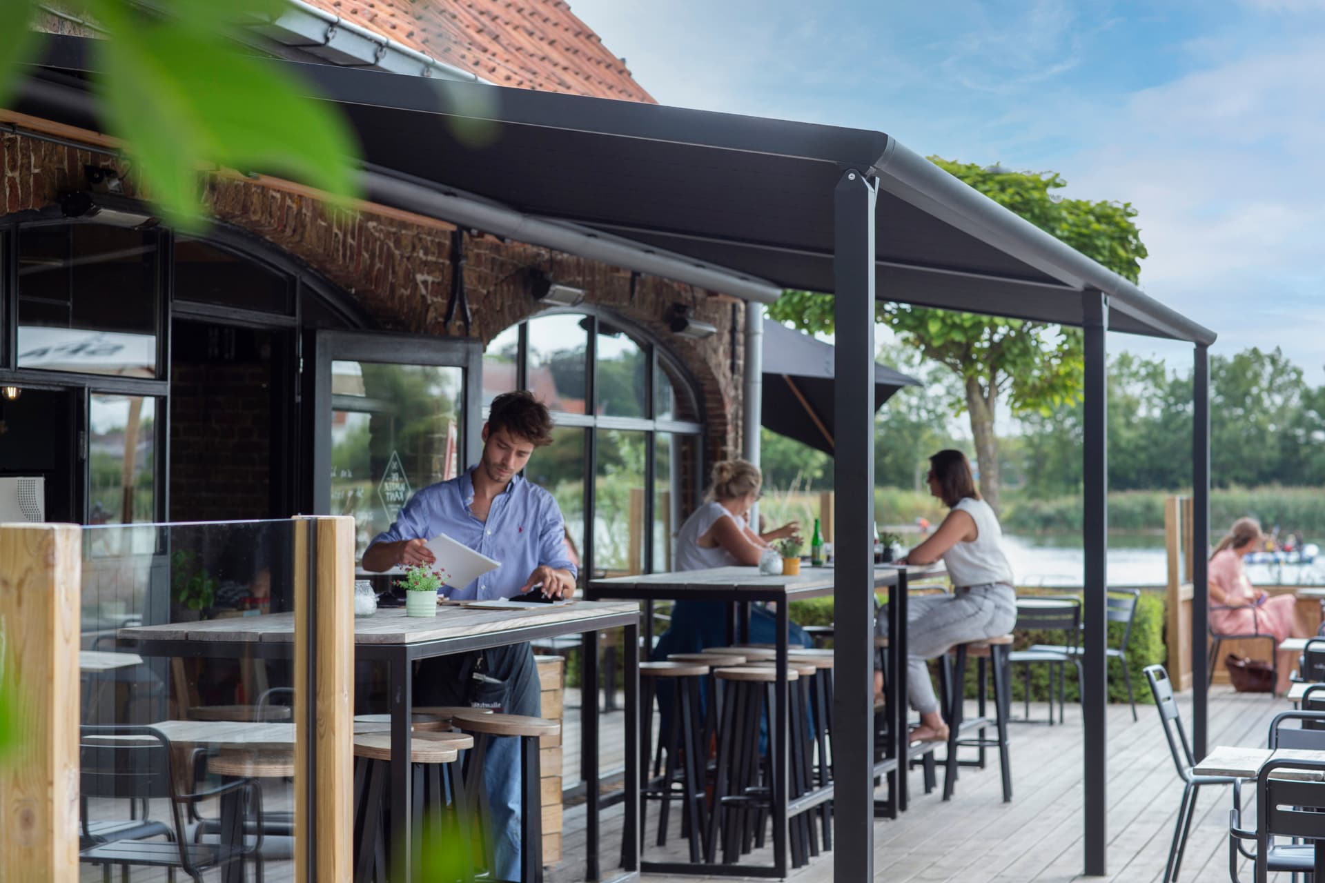 Aluminium pergola‑zonwering die schaduw biedt boven een horecaterras met hoge tafels en zitplaatsen.