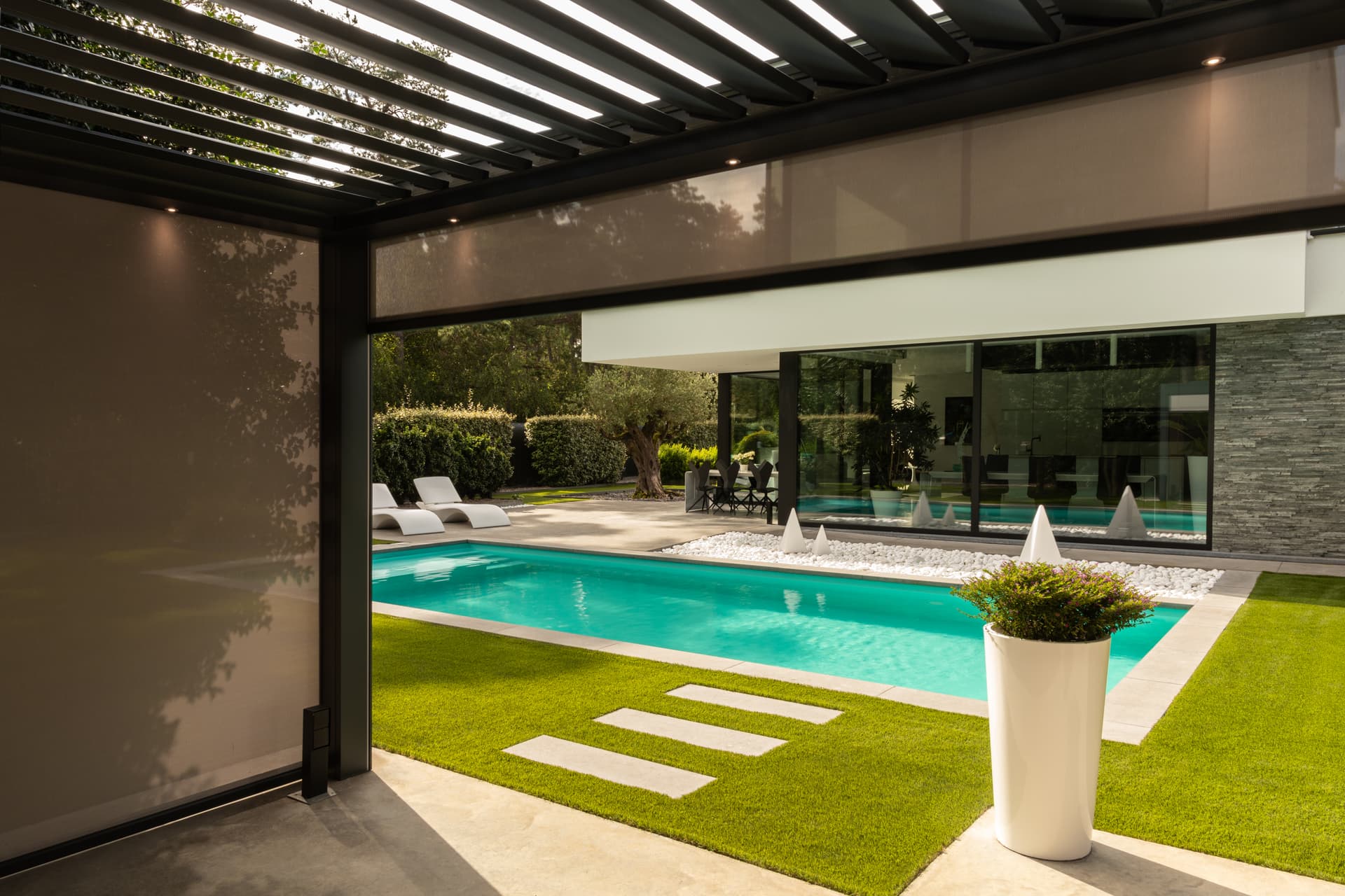 Aluminium pergola met lamellendak en neergelaten ZIP‑screen met zicht op een tuinterras met zwembad en ligstoelen.