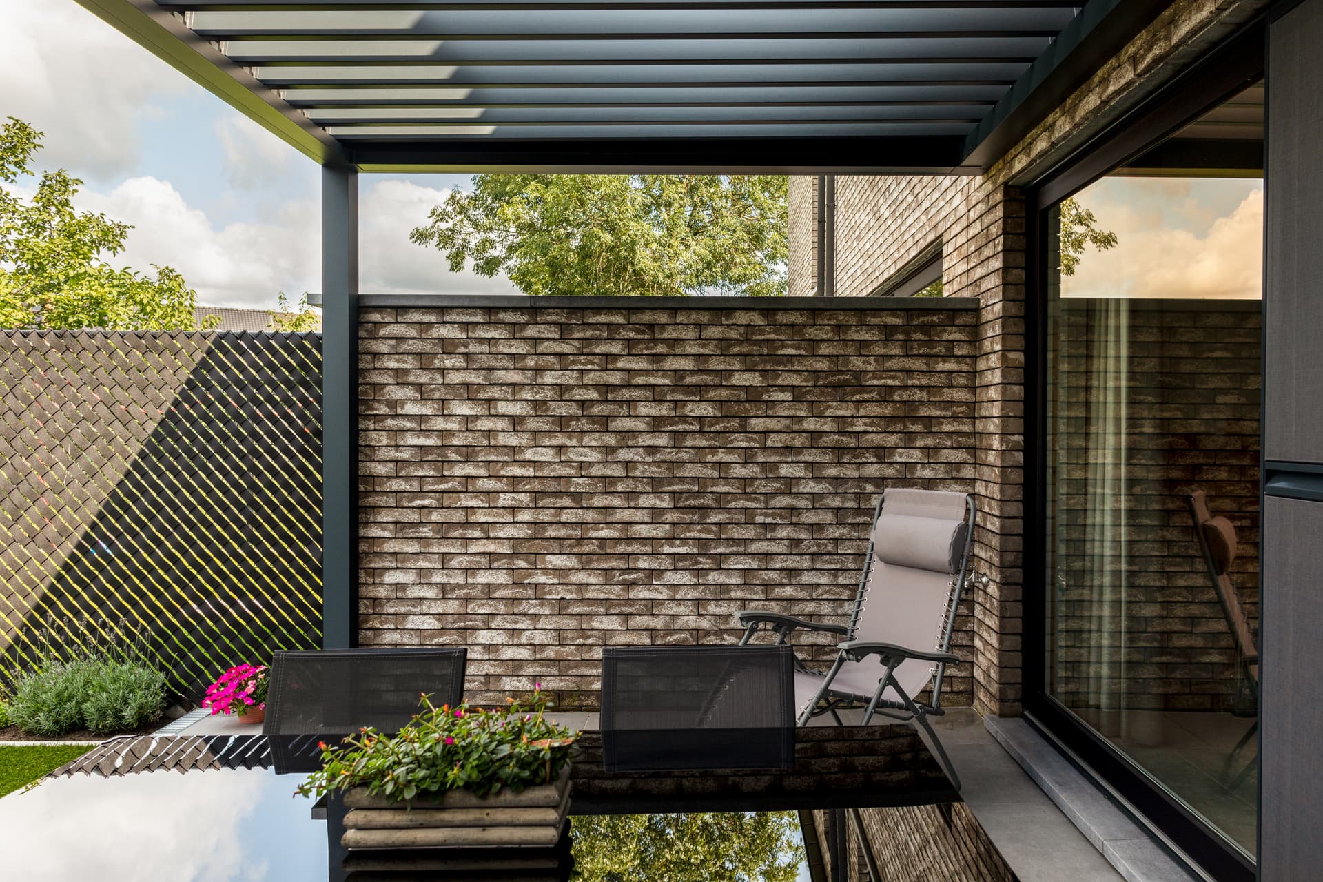 Aluminium pergola met geopend lamellendak dat schuine schaduwpatronen werpt over een terras met buitenzitplaatsen naast een bakstenen gevel.