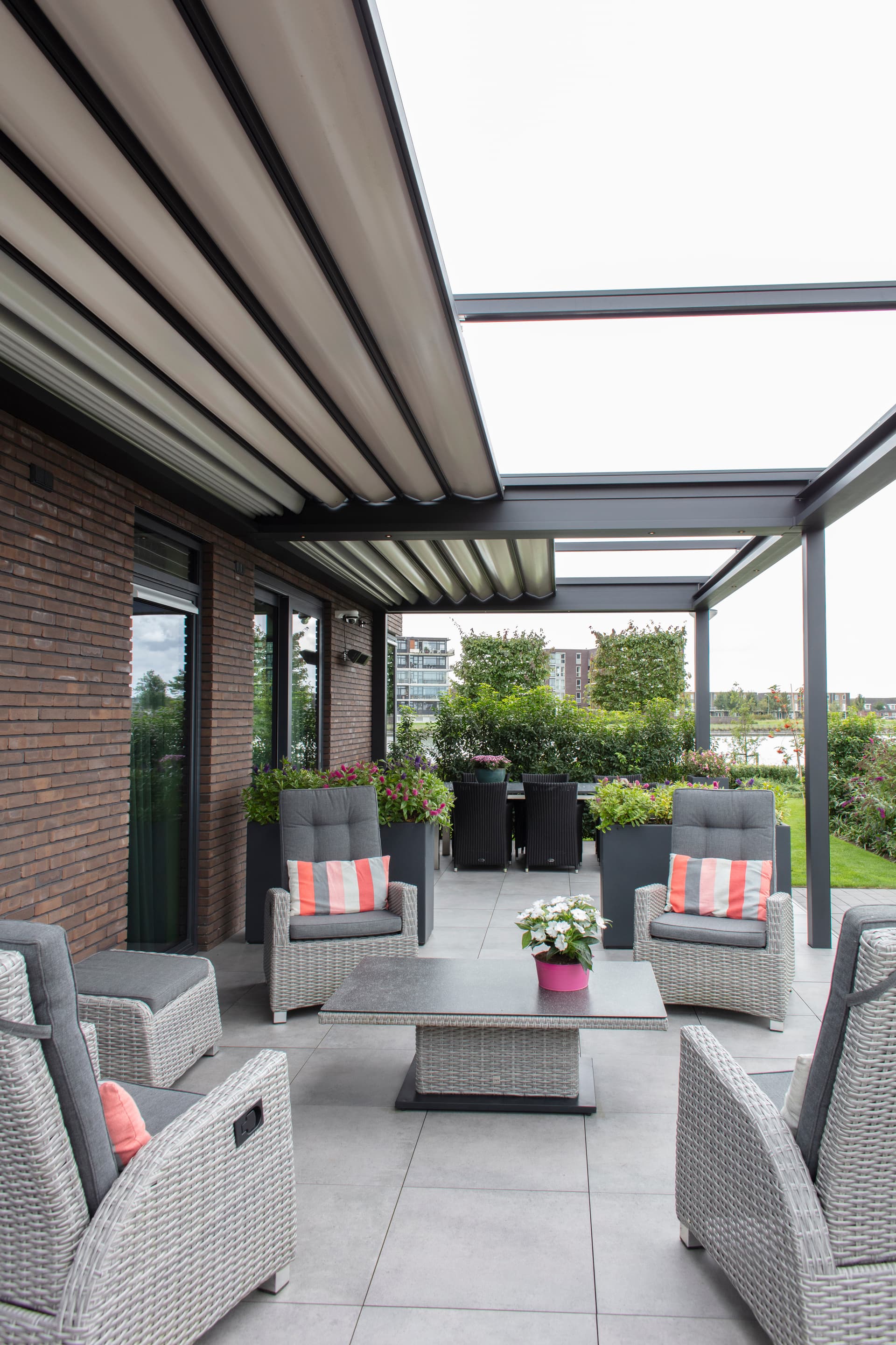 Aluminium B300‑pergola met deels geopend harmonicadoek boven een modern lounge‑ en eetterras omringd door groene tuinbeplanting