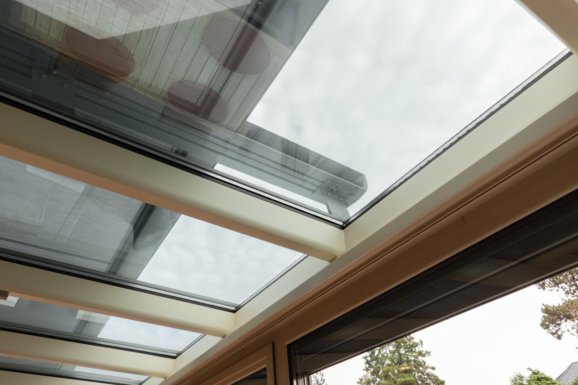 Brustor B129 onder‑glas verandazonwering onder een glazen dak, met strak opgespannen doek en slanke geleiders voor discrete zonwering