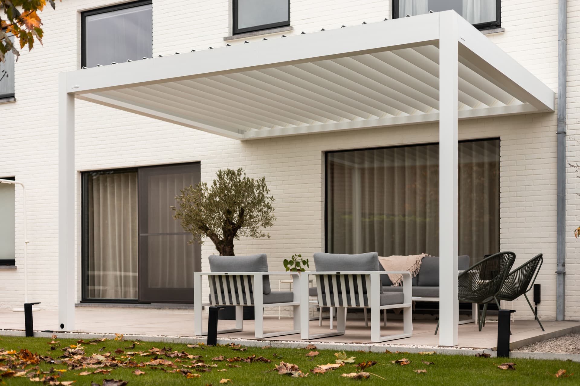 Aluminium pergola met lamellendak die een buitenzitruimte op een terras naast een witte bakstenen woning overschaduwt.