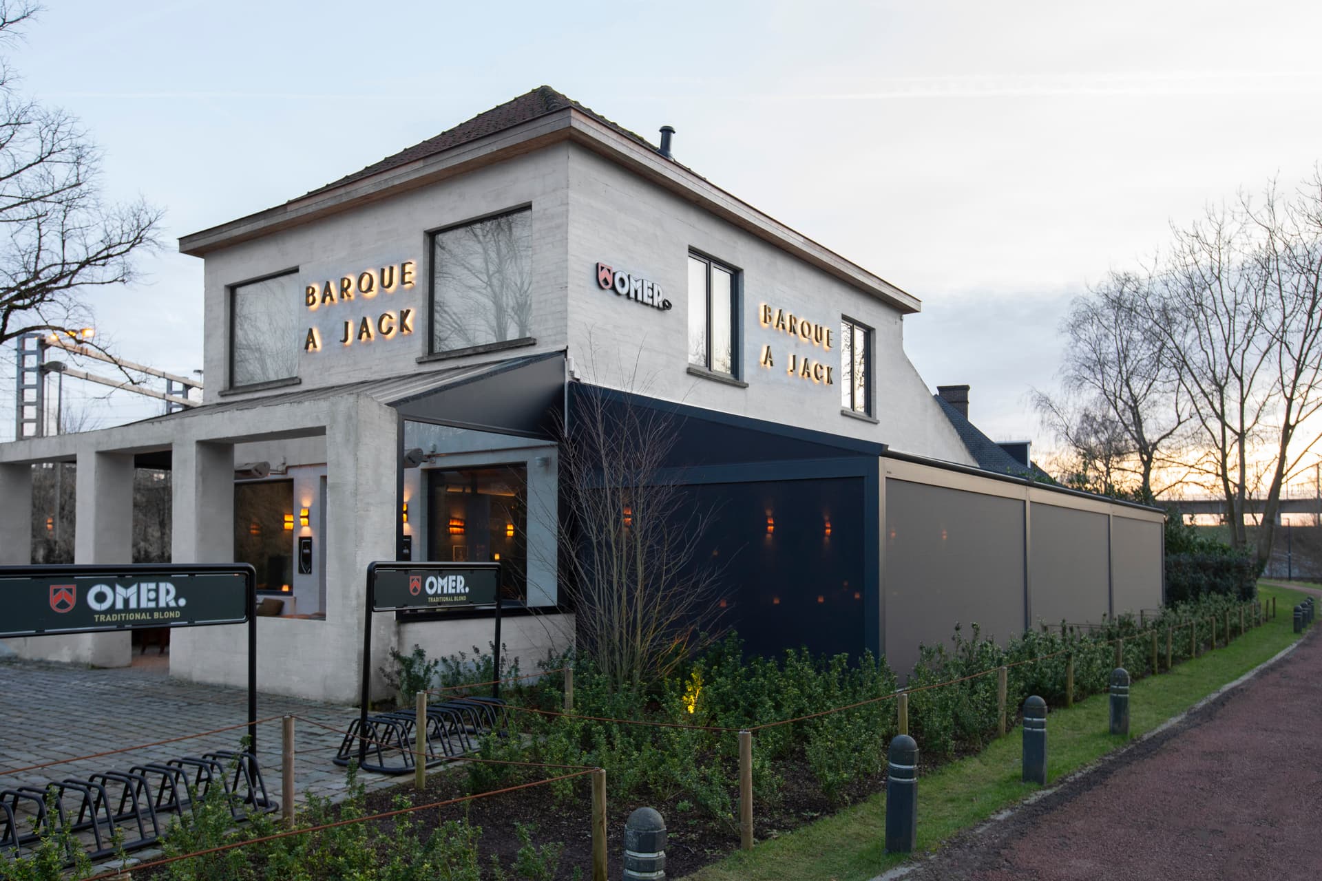 Aluminium B128 XL‑pergola met oprolbaar doekdak geïntegreerd in de gevel van een horecazaak en een warm verlichte, beschutte buitenruimte