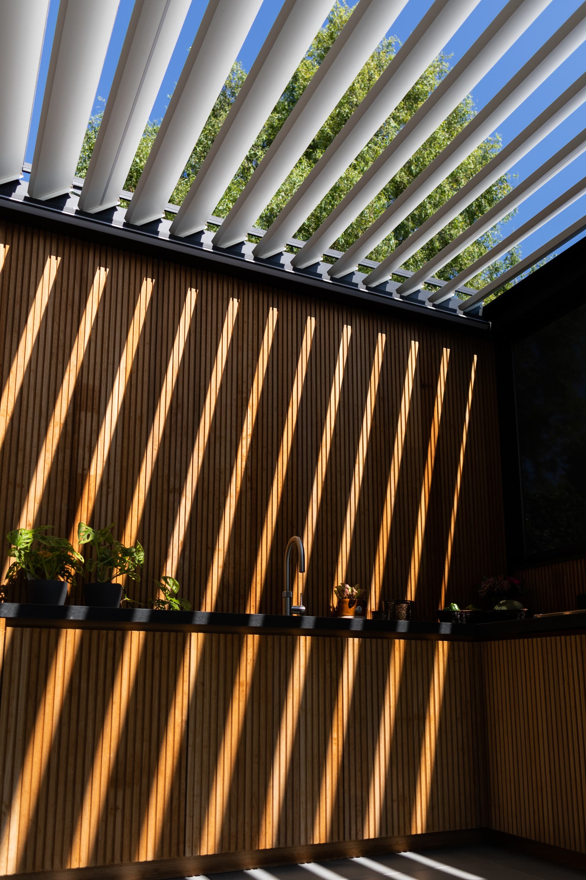 Geopend lamellendak dat een gestreept schaduwpatroon werpt op een houten wand en het werkblad van een buitenkeuken onder de pergola.