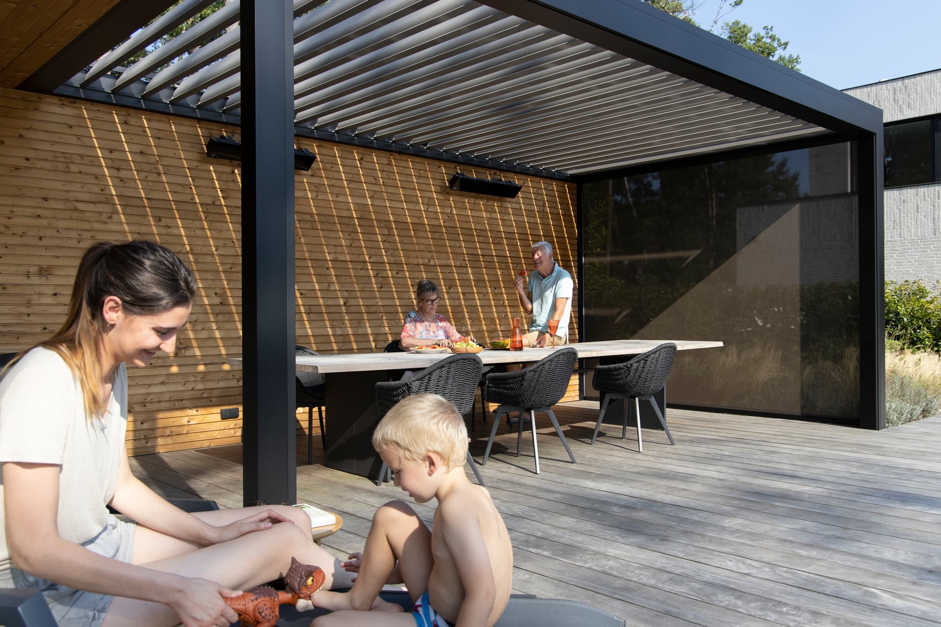 Gezin op een terras onder een aluminium pergola met lamellendak en houten lattenwand, met een buiteneethoek in gedeeltelijke schaduw.