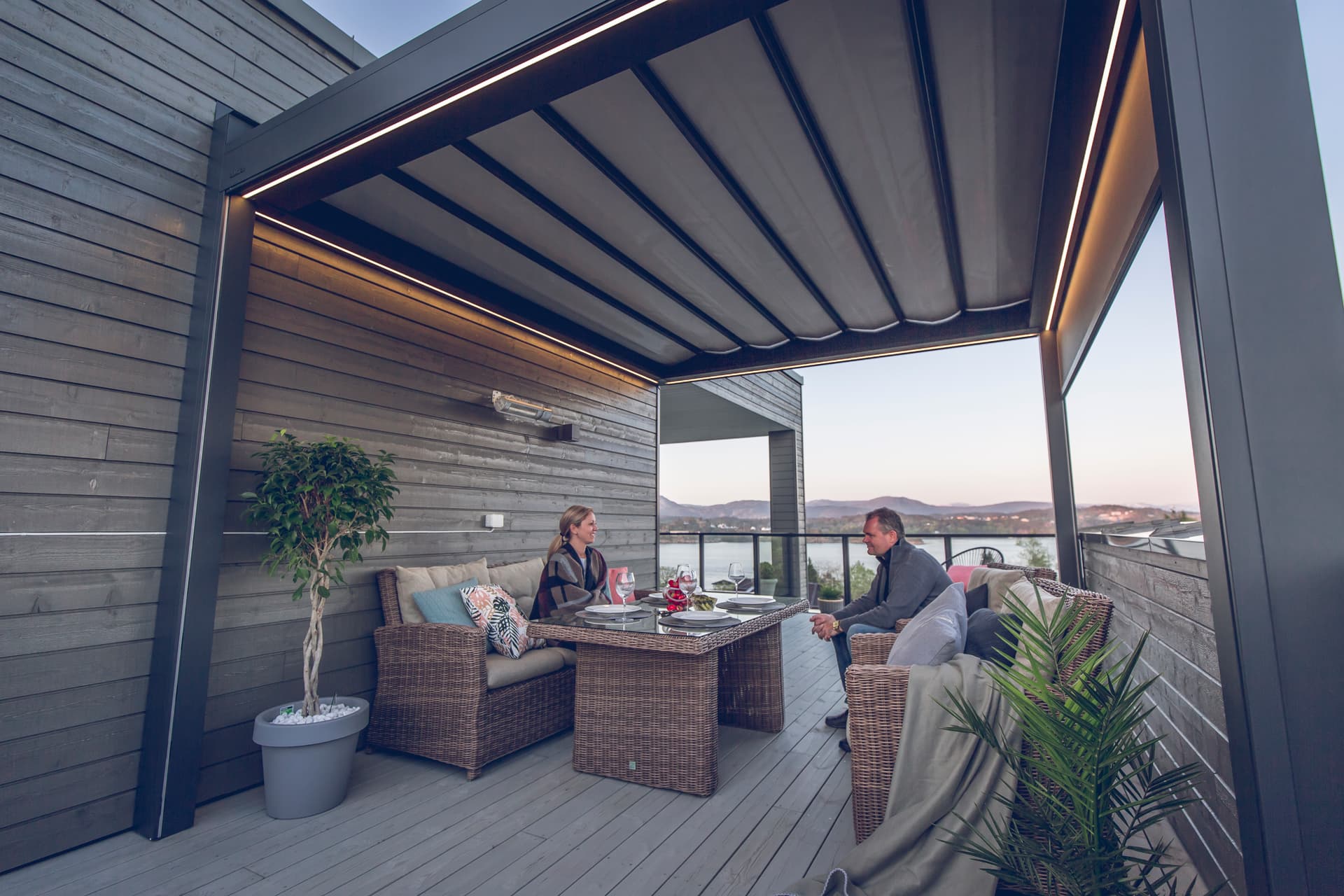 Aluminium B300‑pergola met harmonicadoek en warme geïntegreerde LED‑verlichting boven een gezellige buitenlounge met panoramisch uitzicht