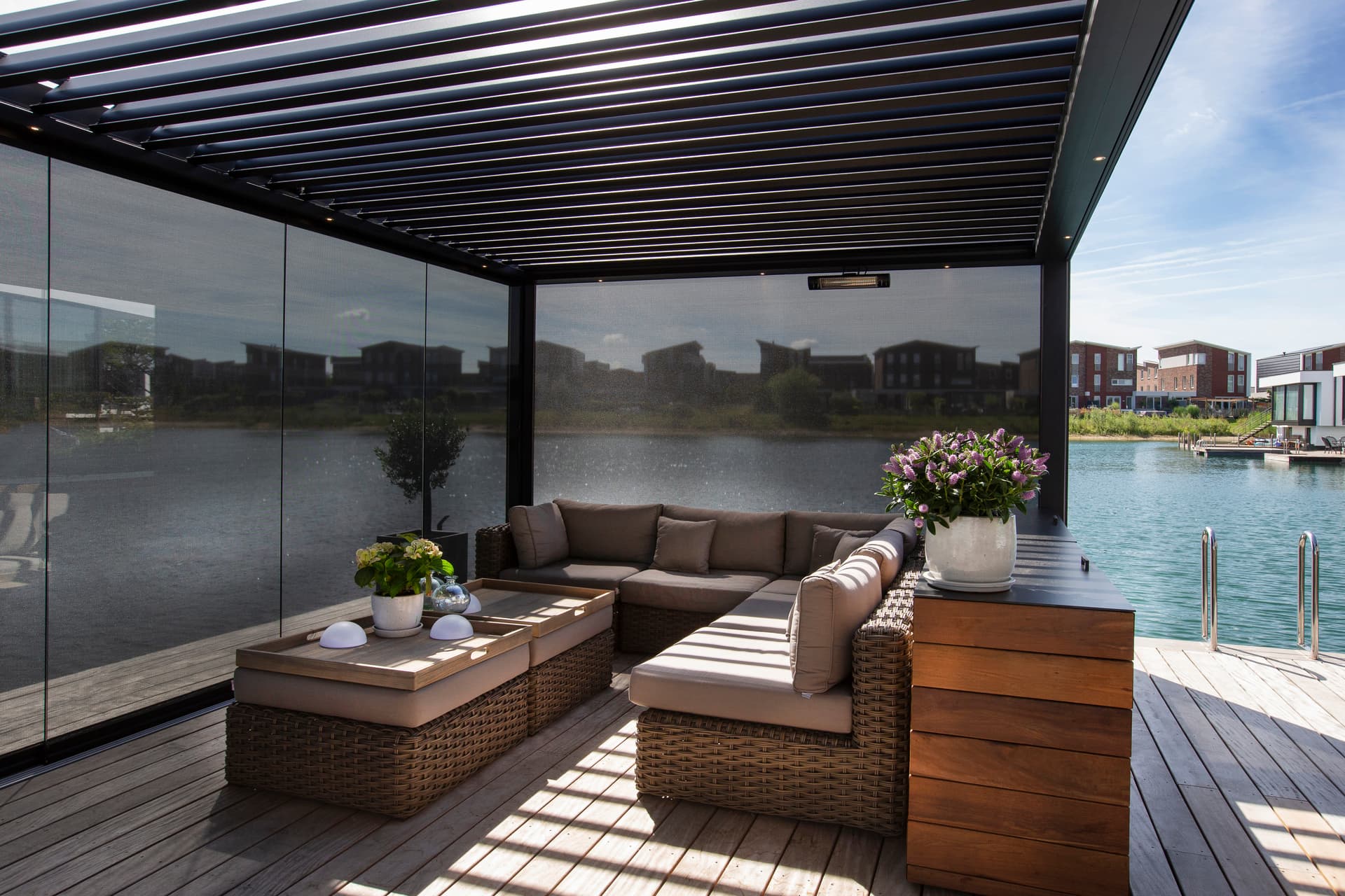 Buitenlounge onder een aluminium pergola met gesloten ZIP‑screen en lamellendak, met uitzicht op het water en een gestreept schaduwpatroon.