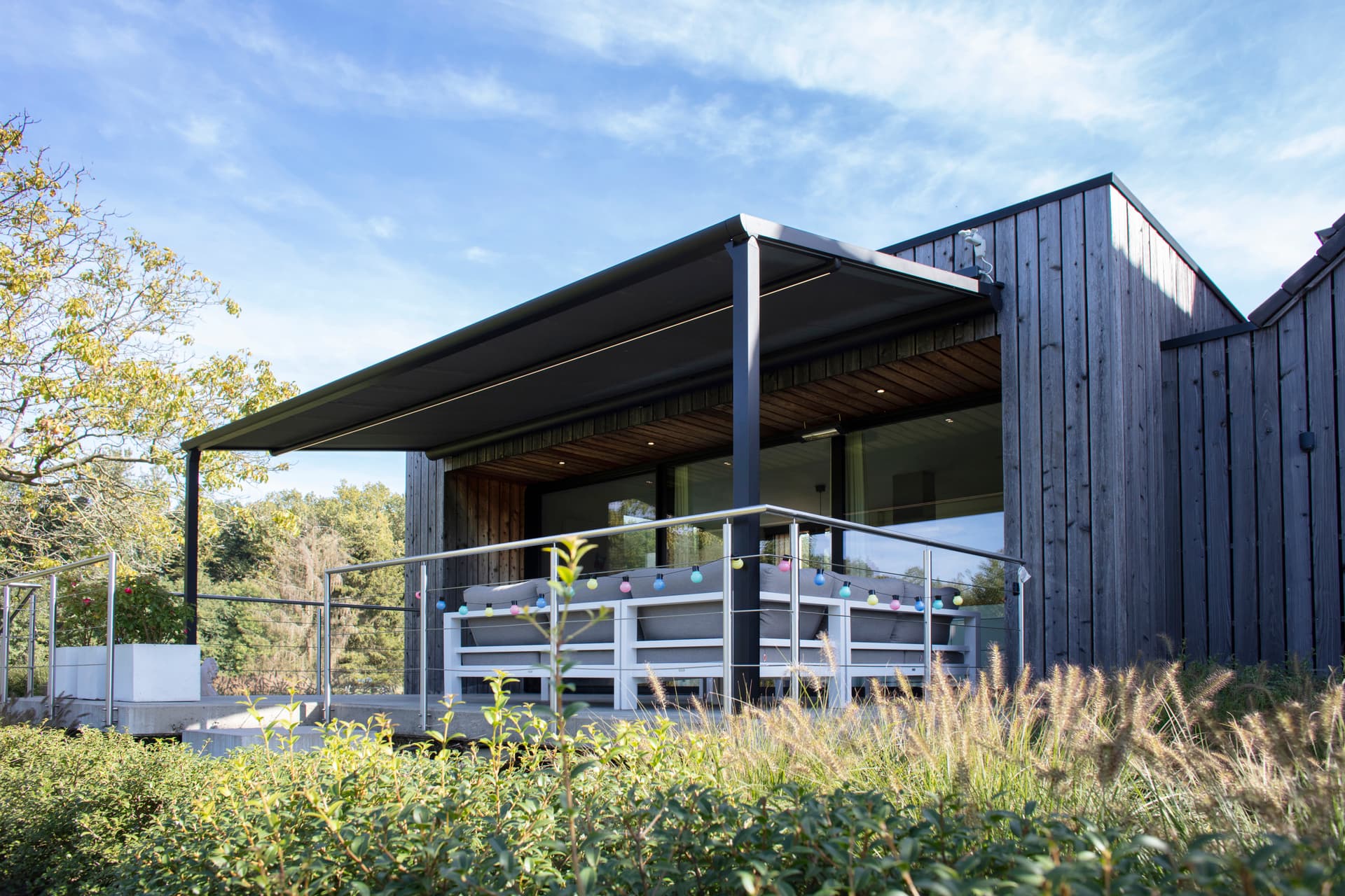 Aluminium pergola met oprolbaar doekdak en geïntegreerde LED‑verlichting boven een moderne loungehoek aan een houten woning
