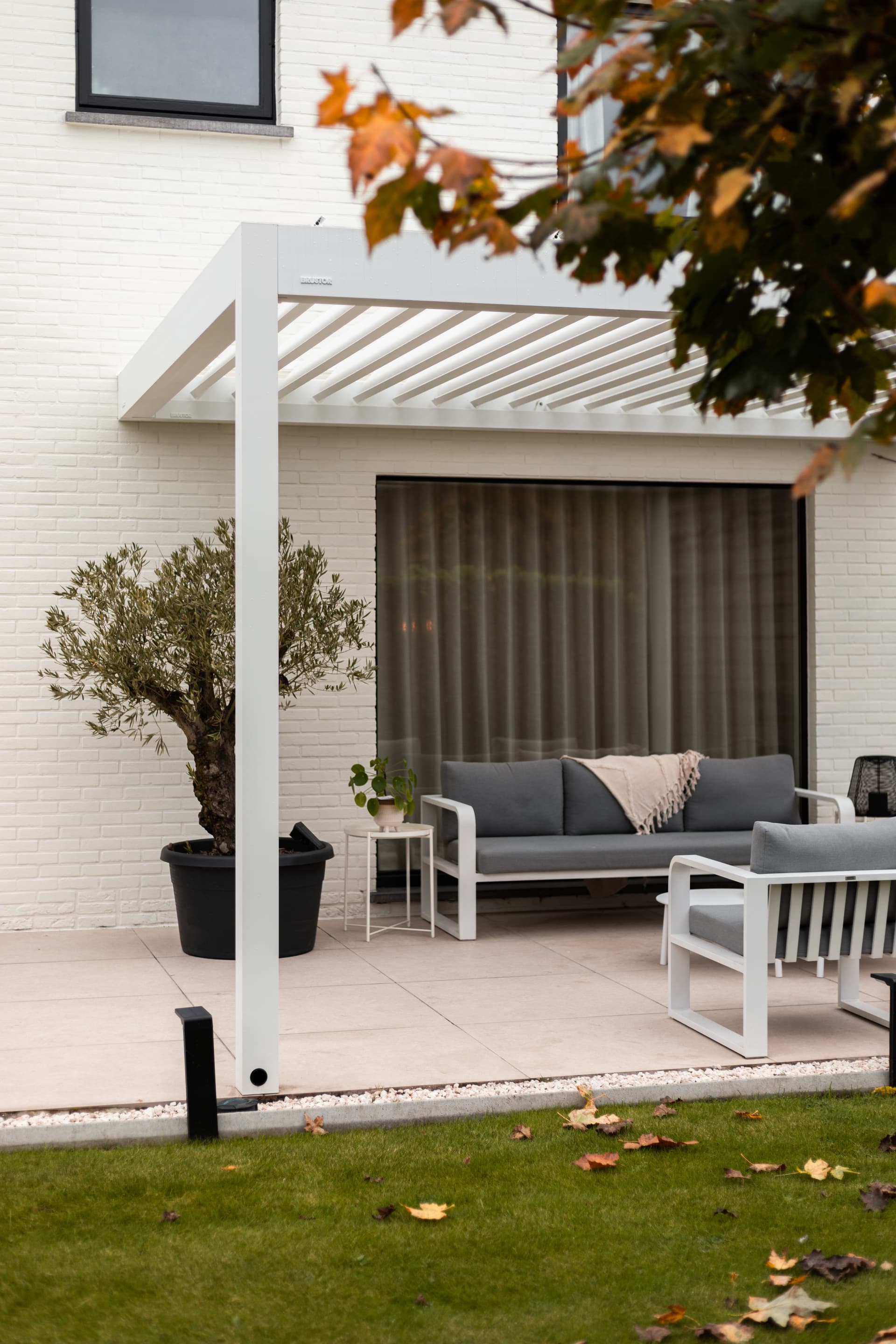 Aluminium pergola met lamellendak op een terras naast een wit bakstenen huis, die een buitenzitruimte overschaduwt.