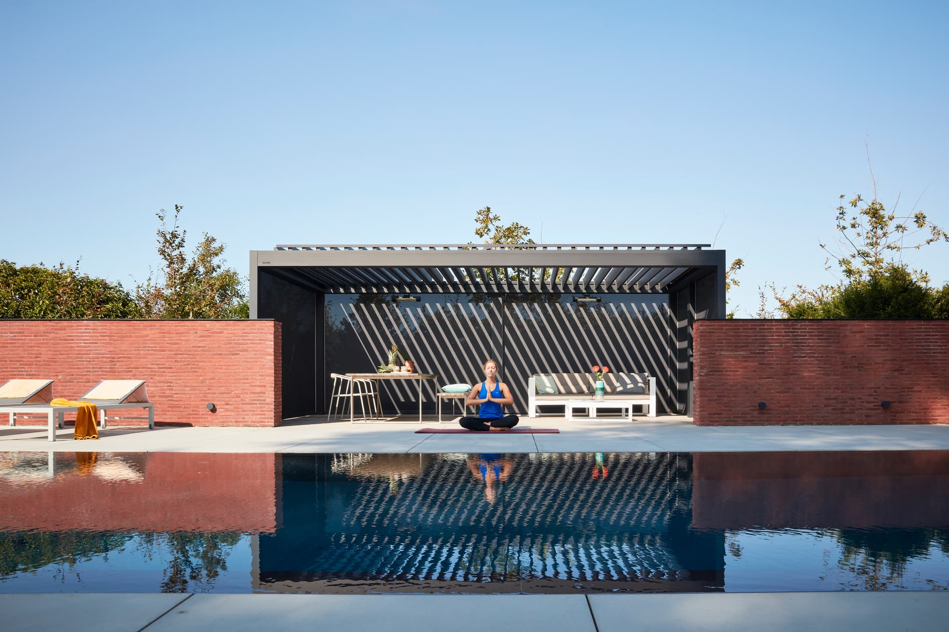 Aluminium pergola met geopend lamellendak dat een gestreept schaduwpatroon werpt over een buitenlounge, met iemand die yoga beoefent op het terras bij het zwembad.