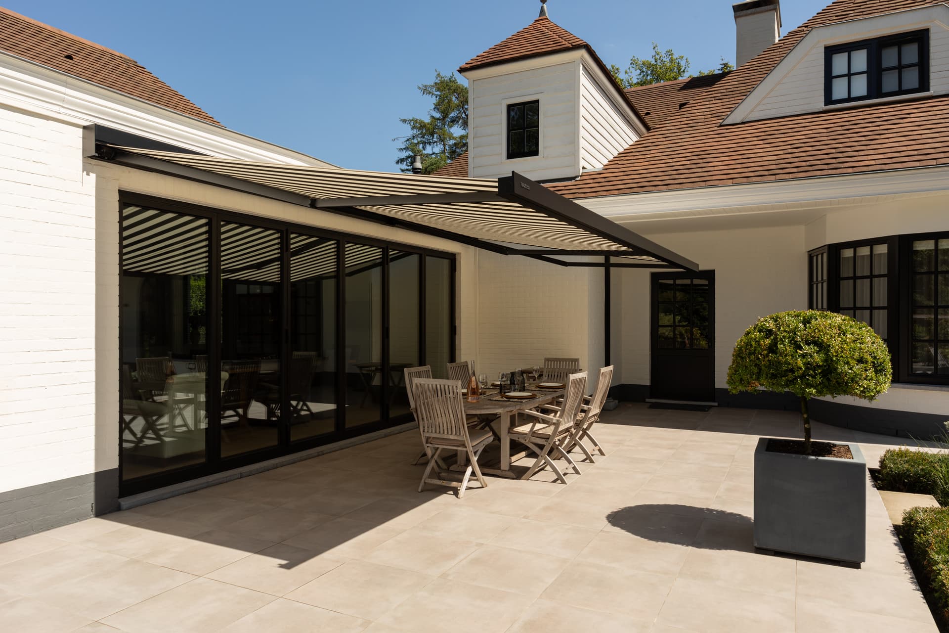 Large retractable awning providing shade over a spacious outdoor dining terrace beside a classic home with wide sliding glass doors
