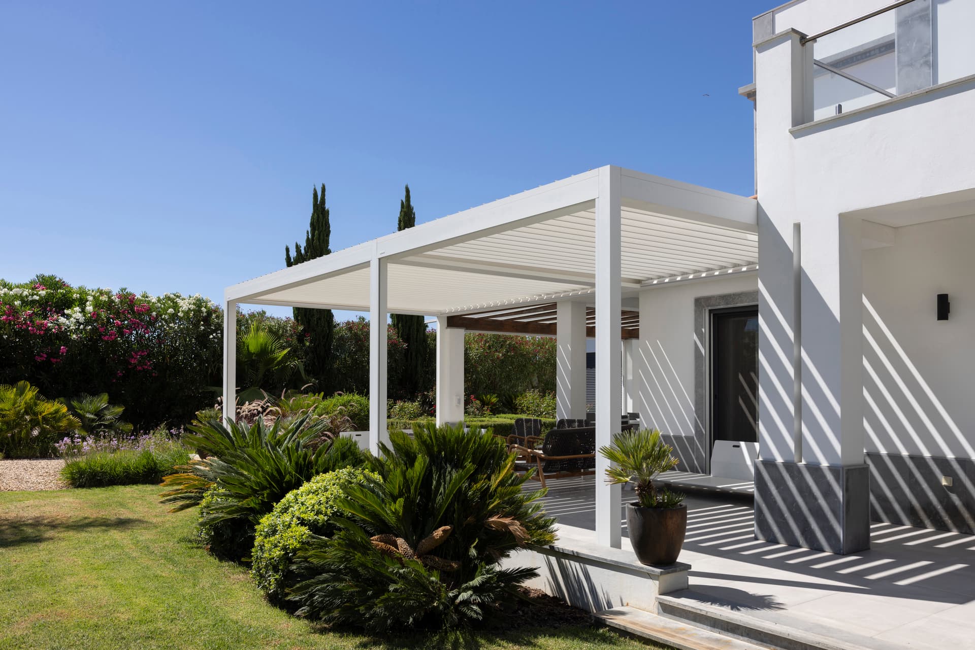 Witte aluminium pergola met lamellendak die een buitenzitruimte op een terras naast een moderne woning overschaduwt, omringd door een aangelegde tuin.