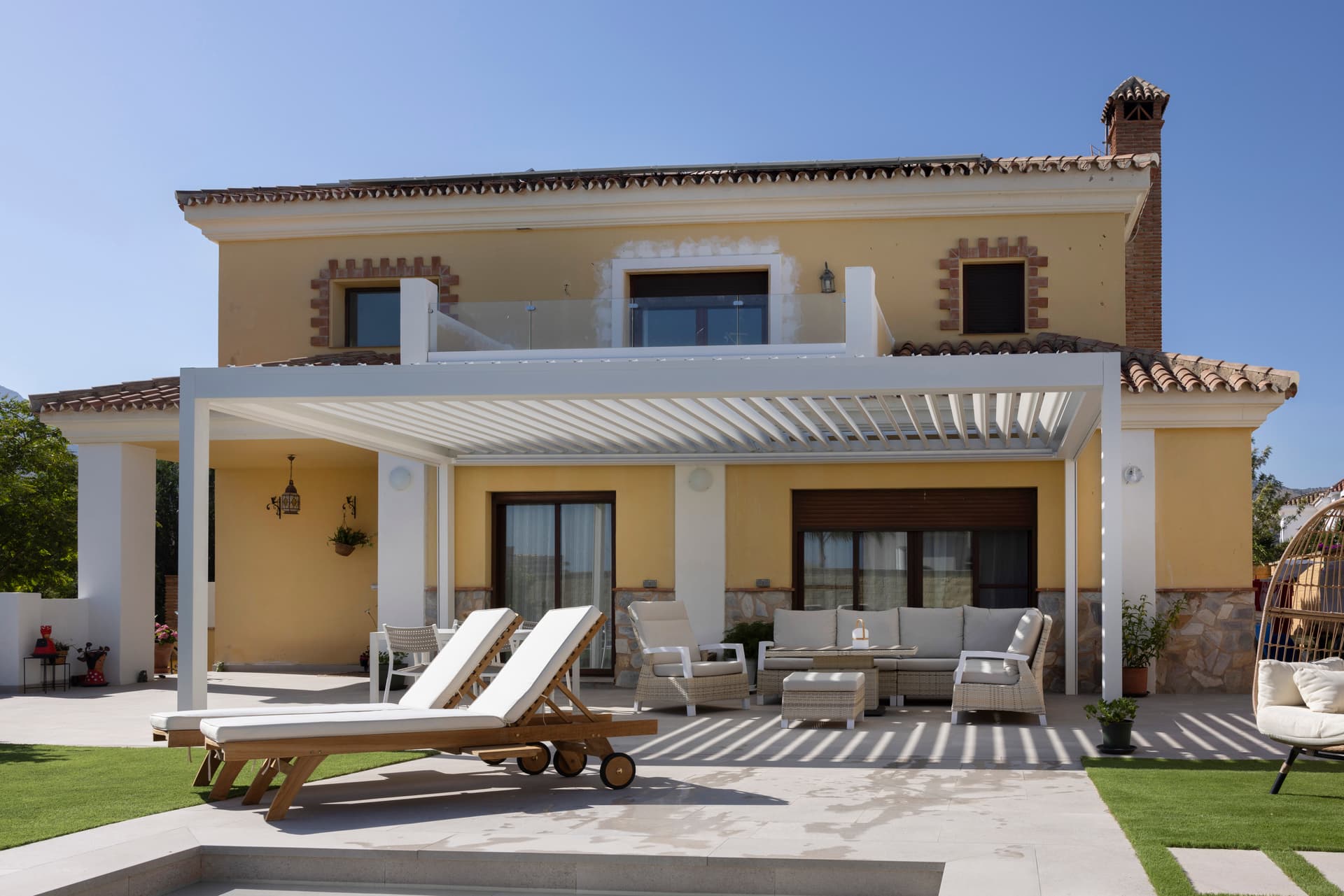 Aluminium pergola met lamellendak die een buitenterras met loungehoek en ligstoelen naast een moderne woning overschaduwt.