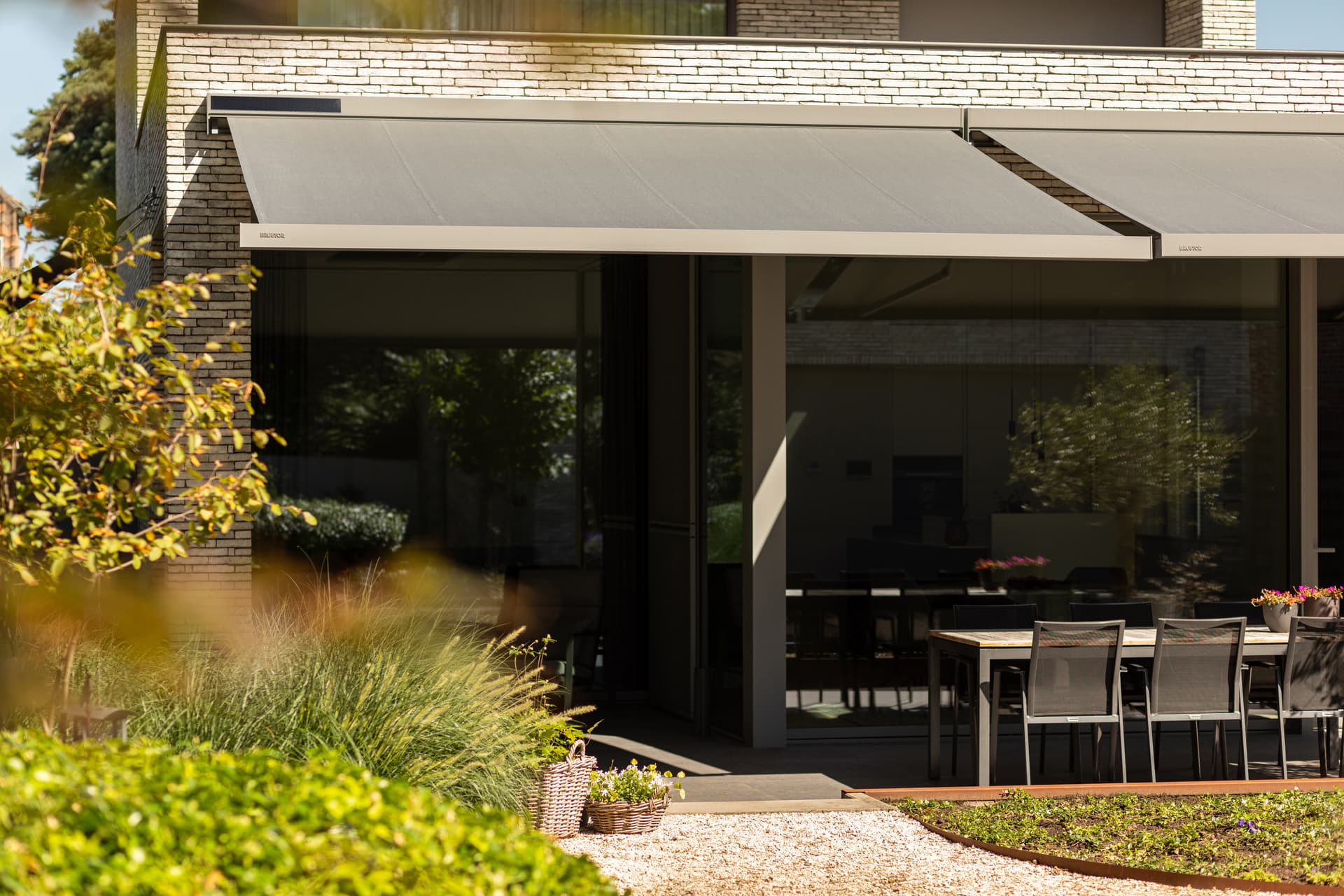 Aluminium retractable awnings in dark finish with integrated solar panel installed above large windows of contemporary home with terrace dining area