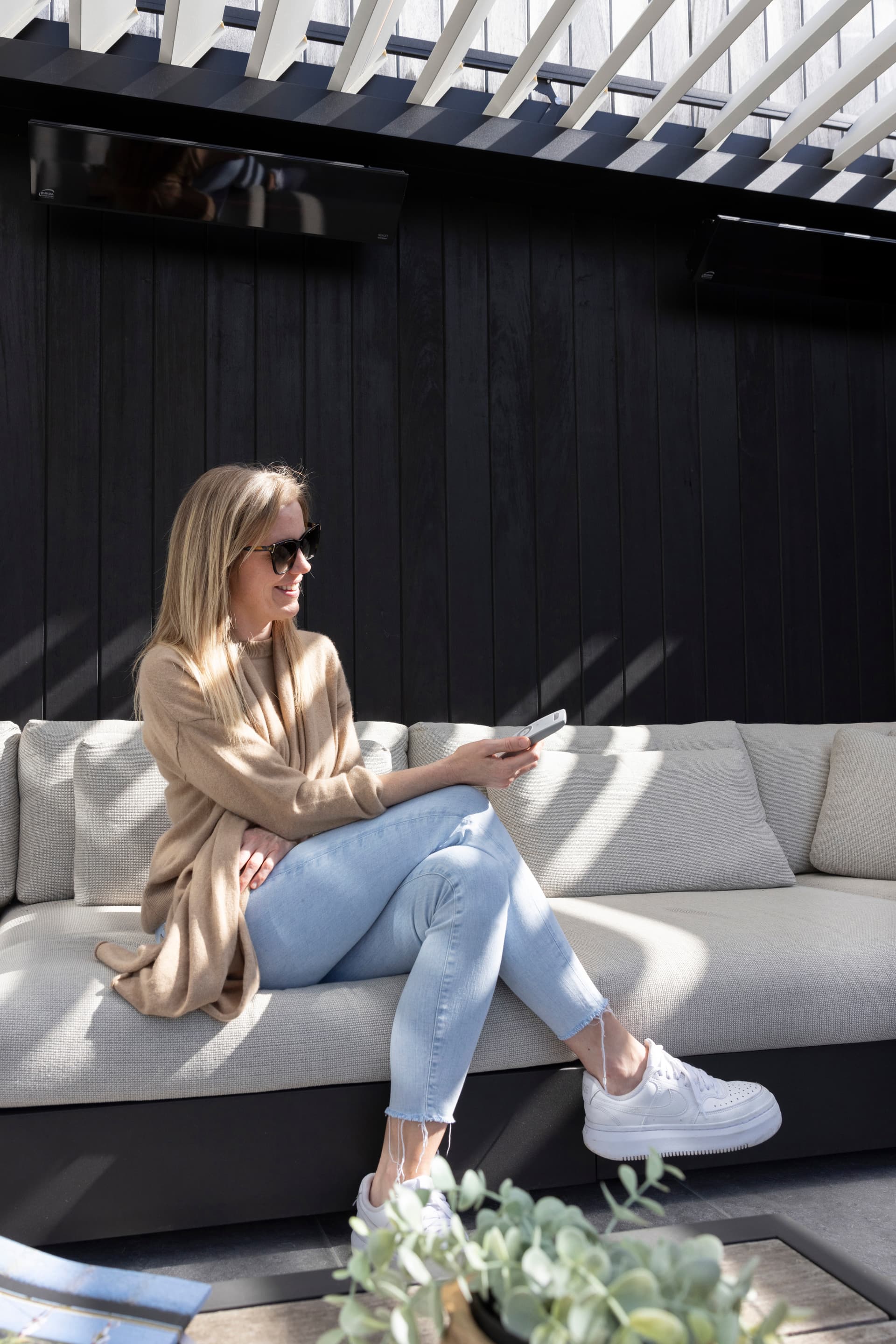 Persoon zittend op een buitensofa onder een pergola met lamellendak, met een afstandsbediening in de hand en een gestreept schaduwpatroon op de zithoek.