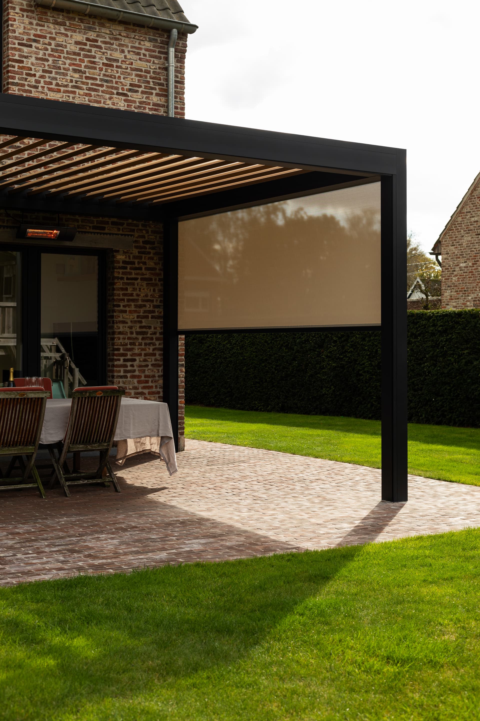 Aluminium pergola met lamellendak en neergelaten ZIP‑screen die een buiteneethoek naast een bakstenen woning overschaduwt.