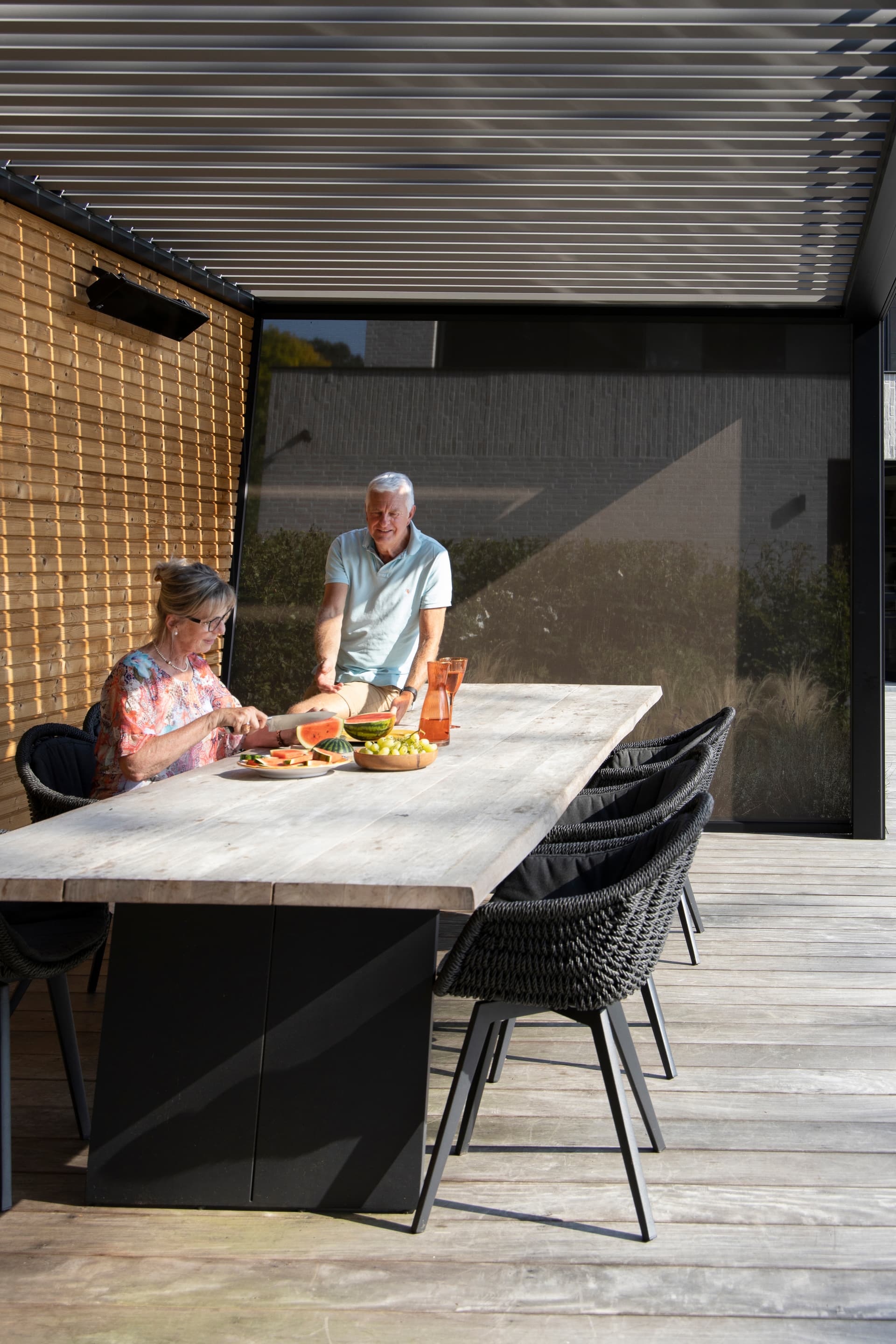 Buiteneethoek onder een aluminium pergola met lamellendak en ZIP‑screen, waar twee personen eten voorbereiden aan een houten tafel op een zonnig terras.