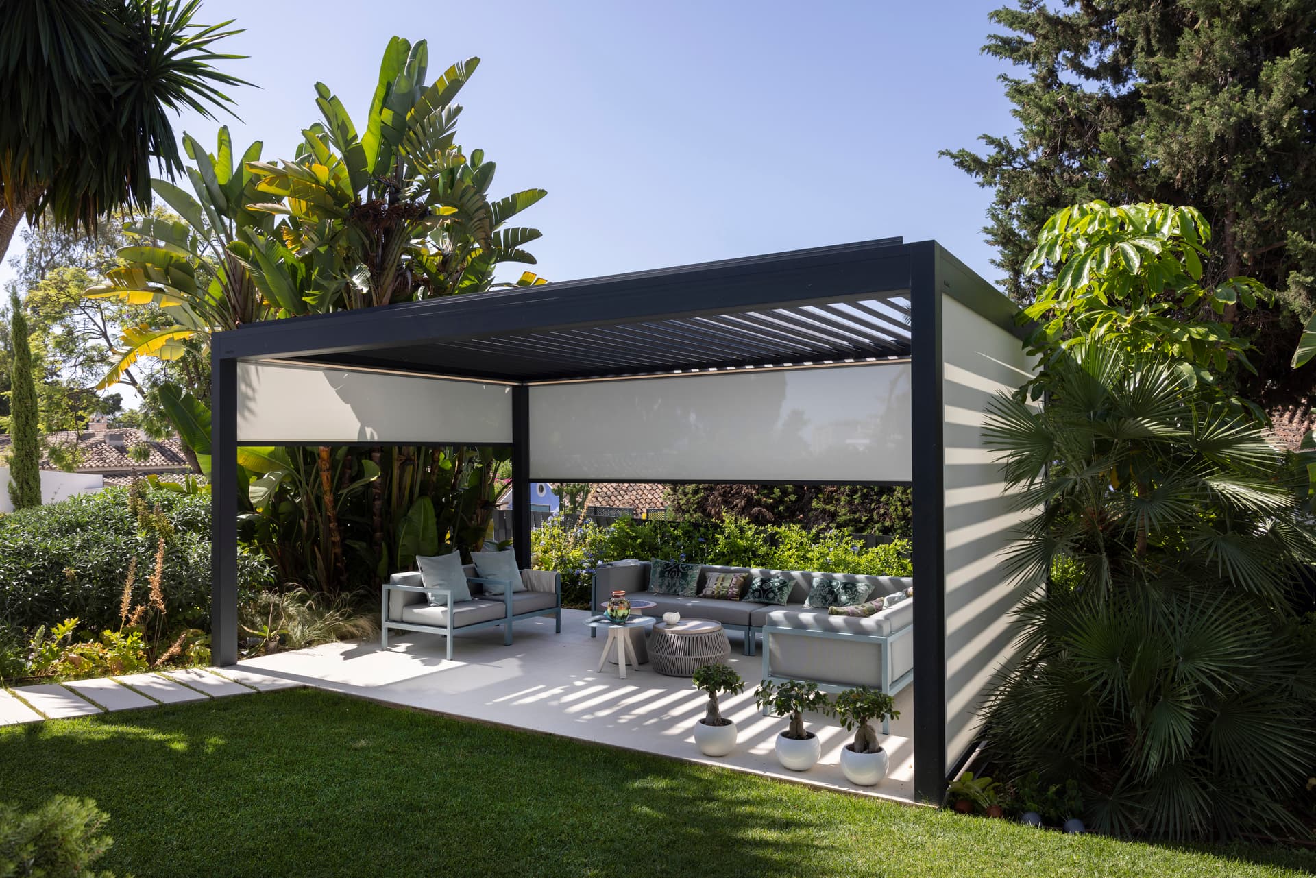 Aluminium pergola with louvred roof and ZIP‑screens enclosing an outdoor lounge area in a landscaped garden with tropical plants.