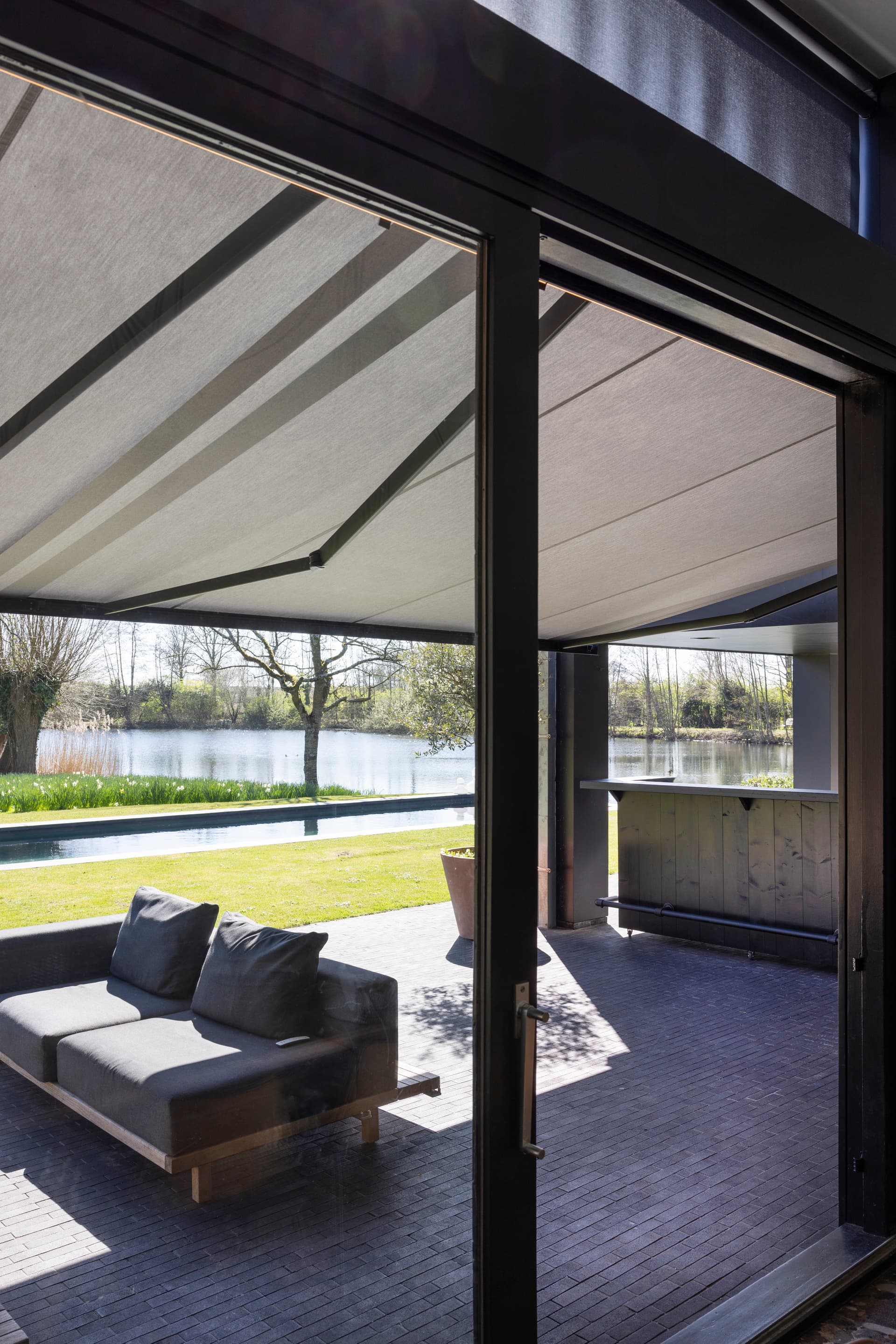 Aluminium retractable awning with light‑coloured fabric installed above terrace with lounge area overlooking garden and water