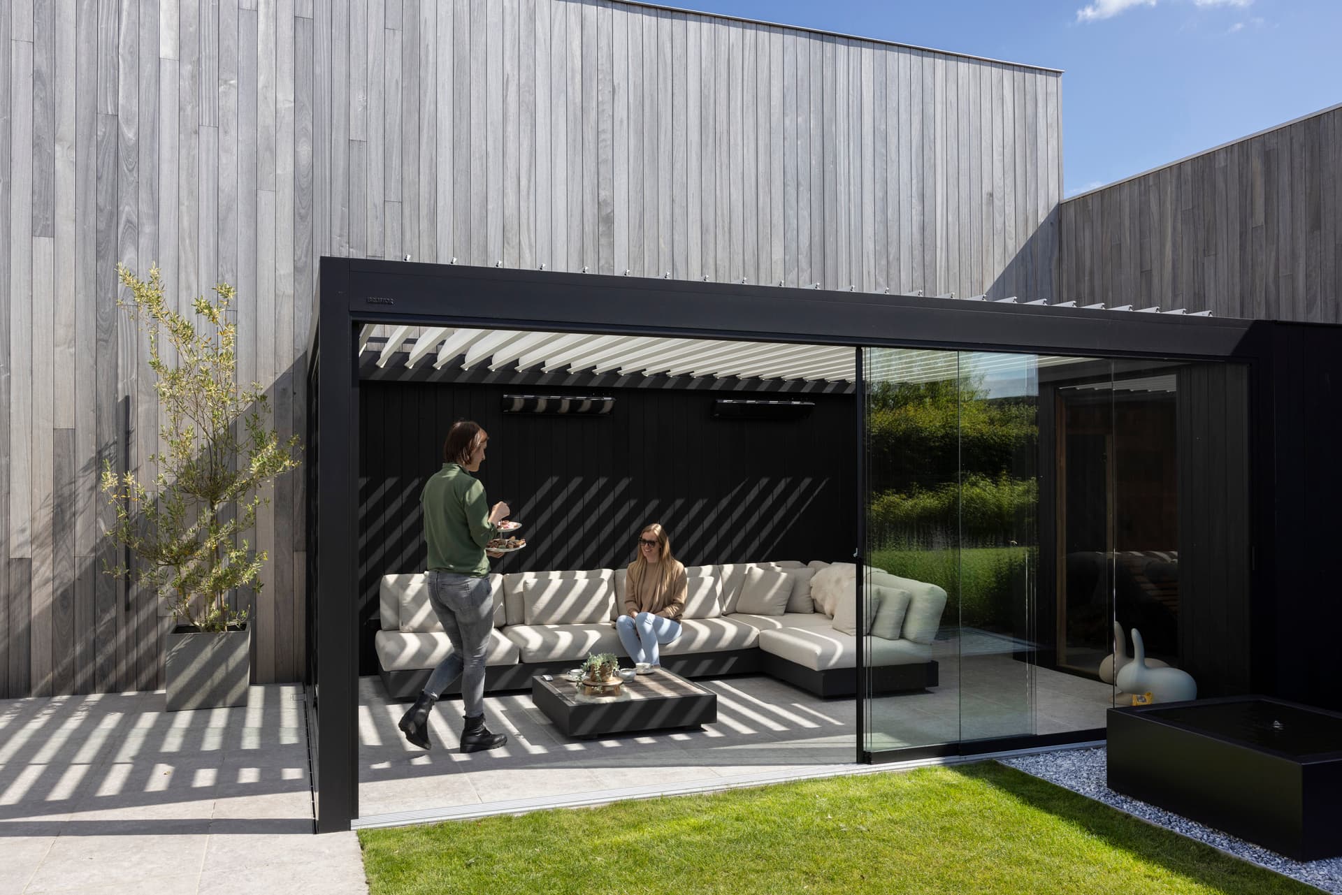 Aluminium pergola met lamellendak en glazen schuifwanden rond een buitenlounge, met een gestreept schaduwpatroon op het terras.