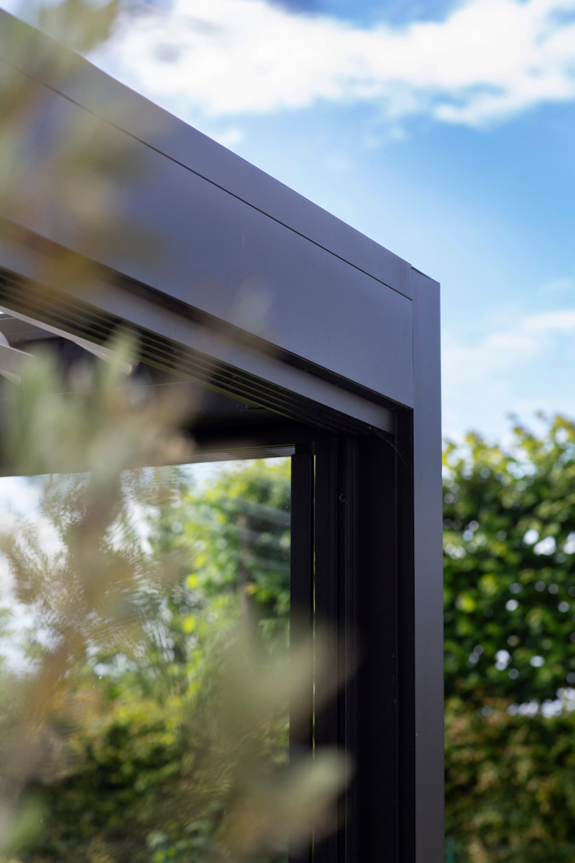 Close‑up van het hoekprofiel van een aluminium pergola, met zicht op de strakke afwerking en tuinbegroeiing op de achtergrond.